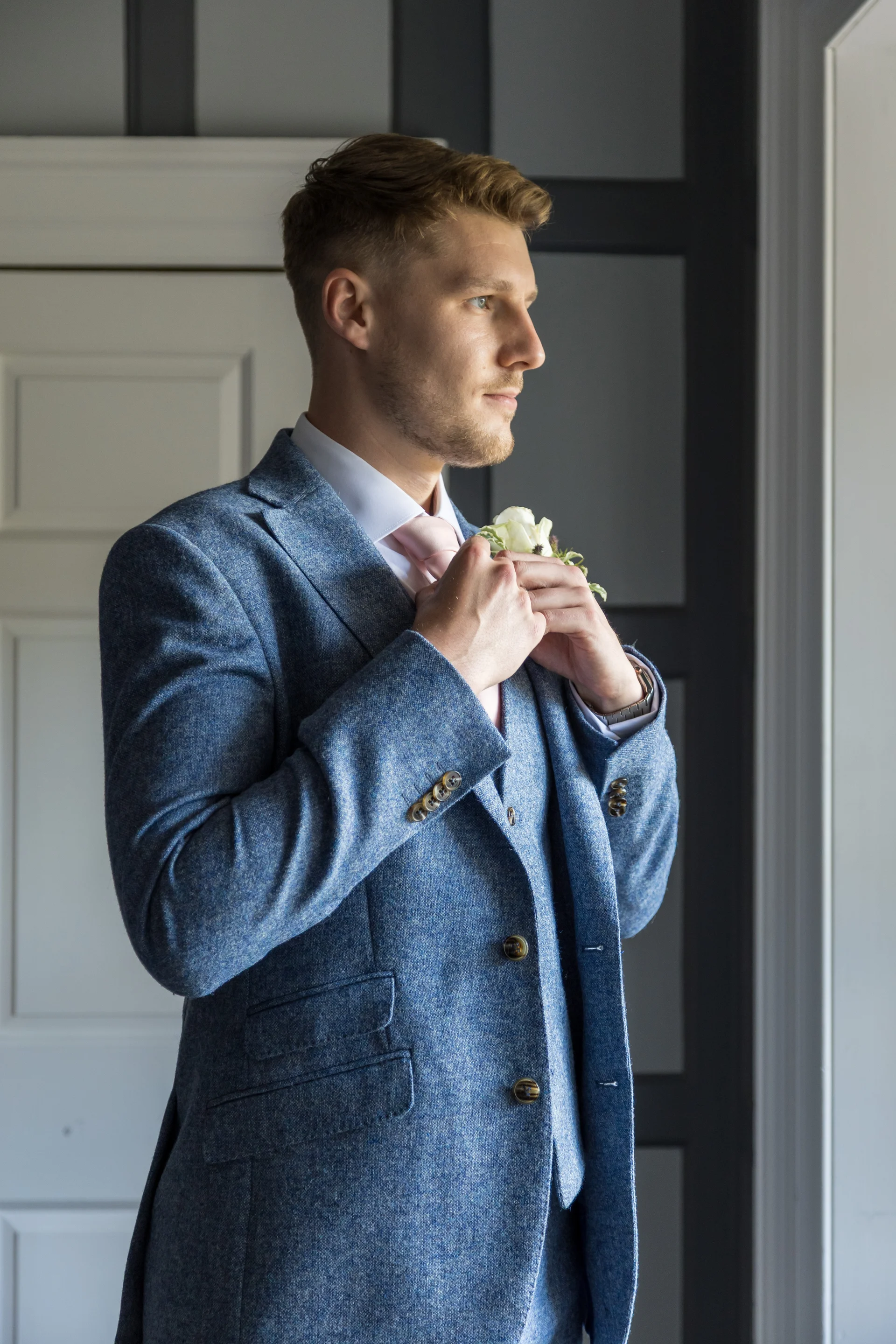 A handsome groom in a textured blue tweed three-piece suit adjusts his pink tie while standing by a bright window during wedding preparations.