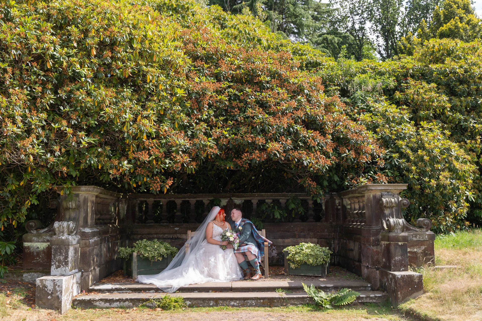 A newlywed couple sits together on a stone bench framed by an ornate balustrade and lush green foliage in a manicured garden setting.