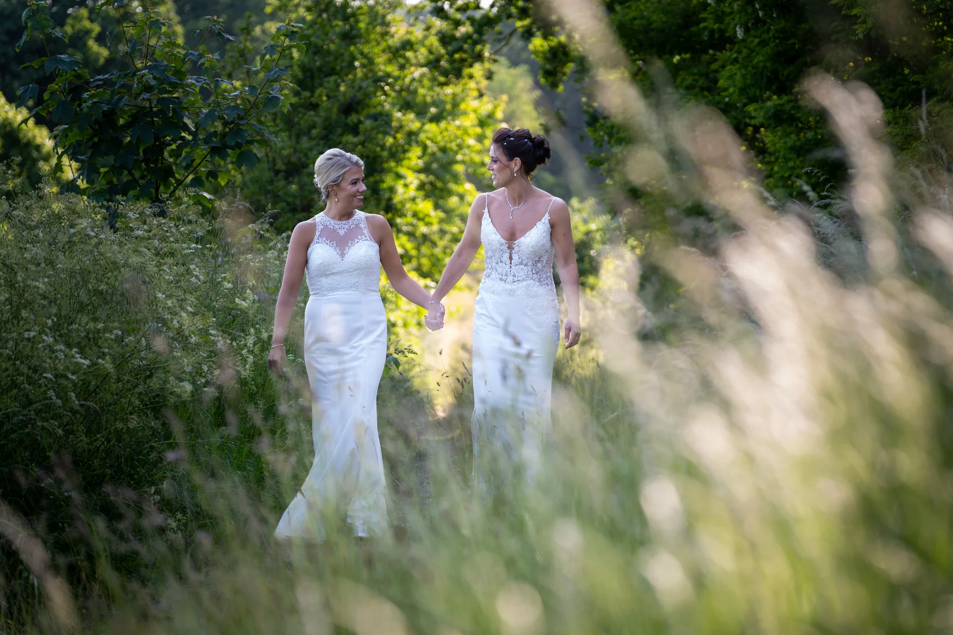 Two smiling brides in white wedding dresses walk hand in hand through a lush green woodland path, framed by tall grass in the foreground.