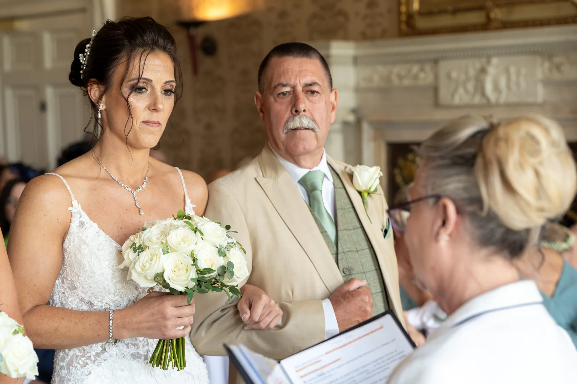 A bride holding a white rose bouquet stands alongside her father in a tan suit as they face the celebrant during a wedding ceremony inside a historic country house.