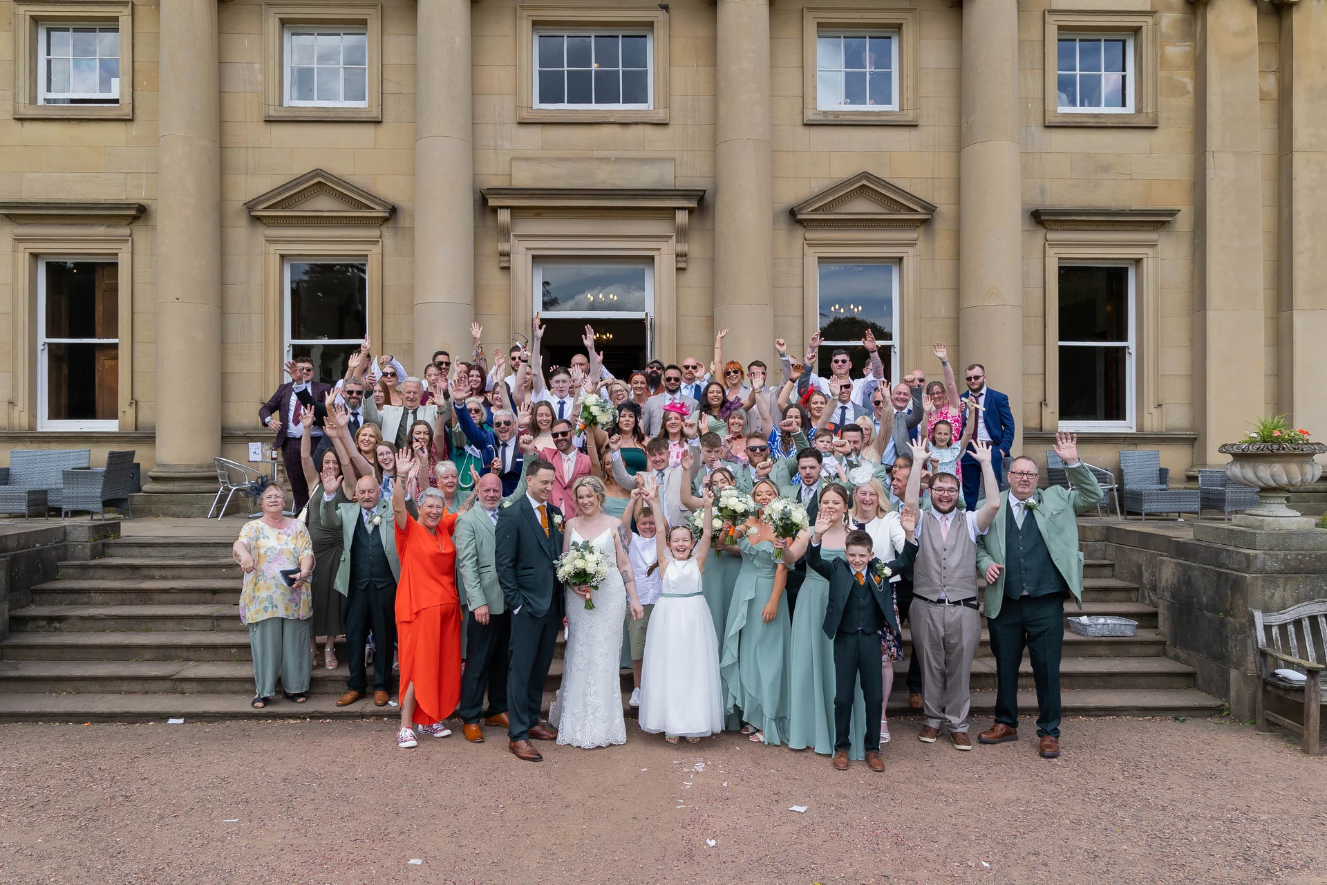 A large wedding party and guests raise their hands in celebration outside a grand stone country house with impressive columns.