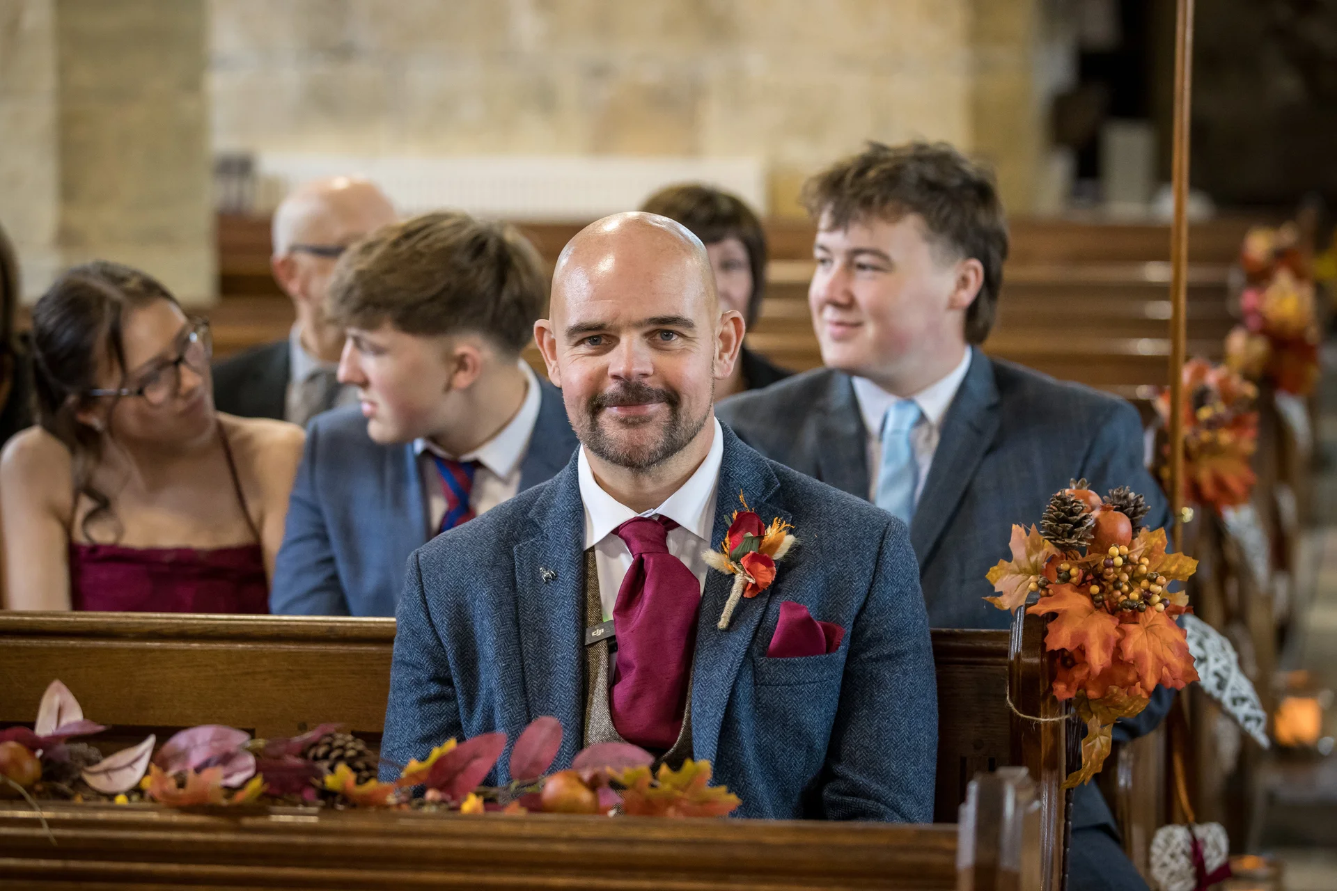 A smiling guest wearing a blue tweed jacket and burgundy tie sits in a wooden church pew decorated with autumn leaves.