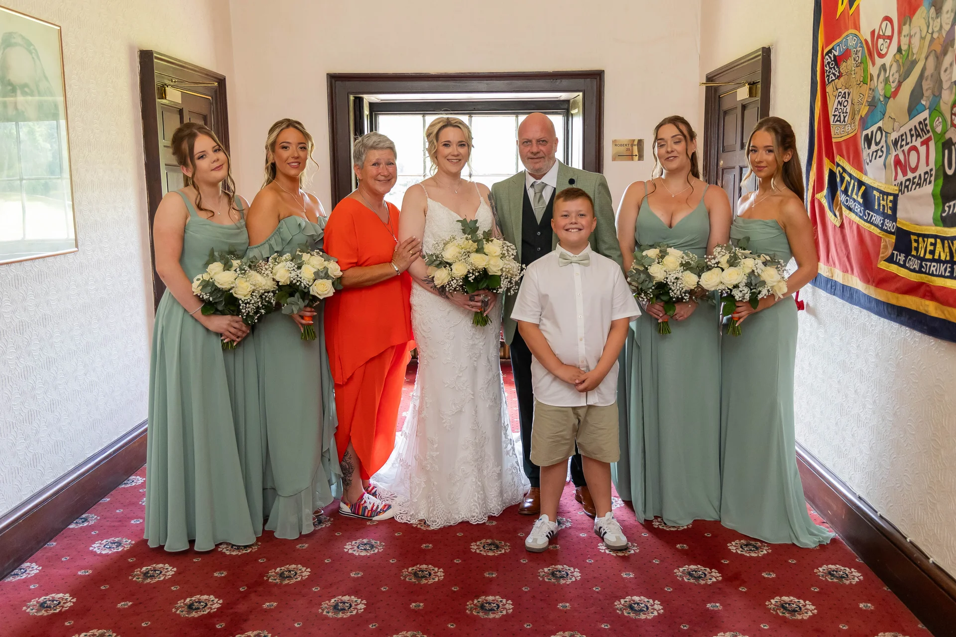 The bride and groom pose happily indoors alongside their bridal party wearing sage green dresses and a young page boy.
