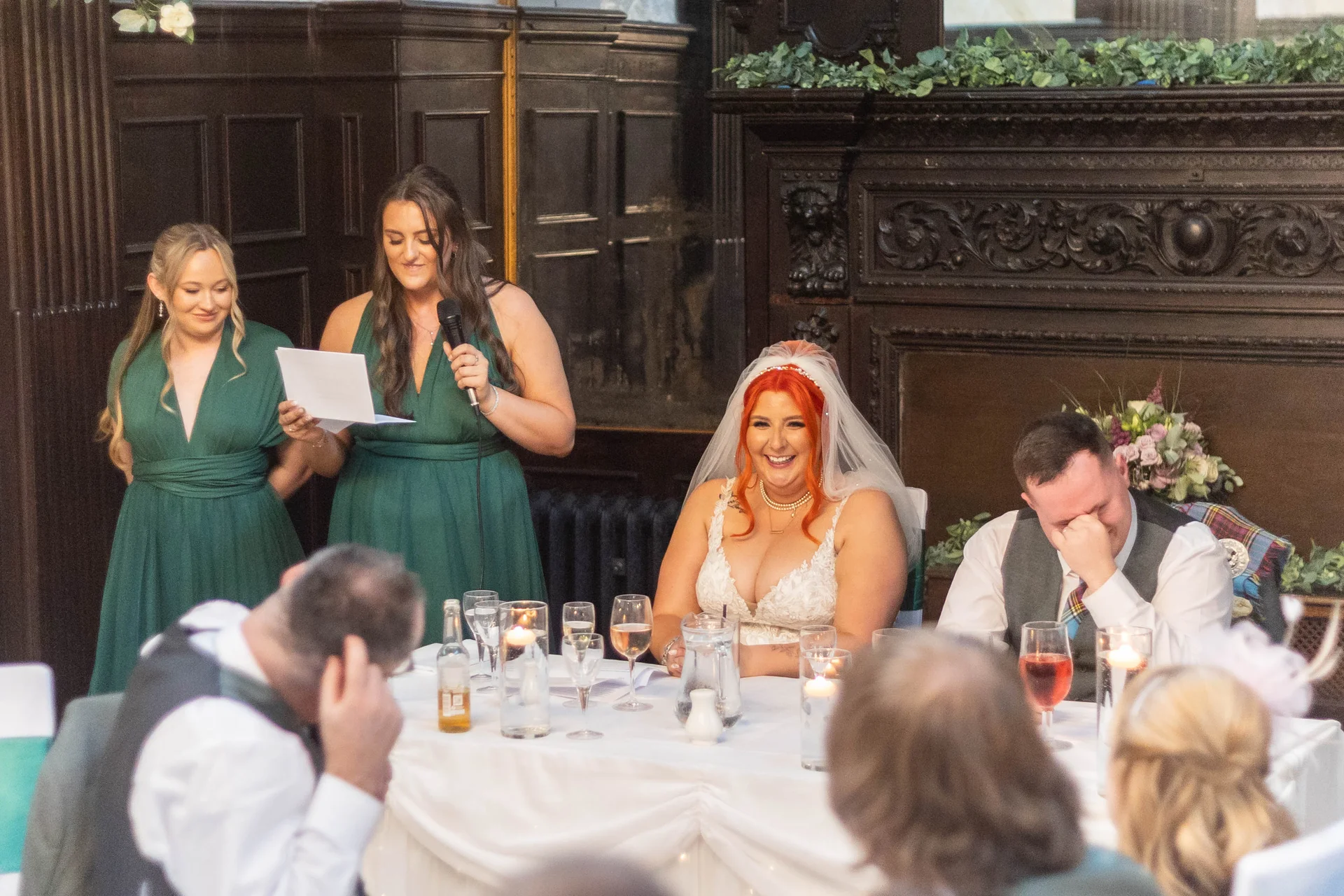 A maid of honour delivers a speech at the top table while the bride laughs and the groom covers his face in amusement.