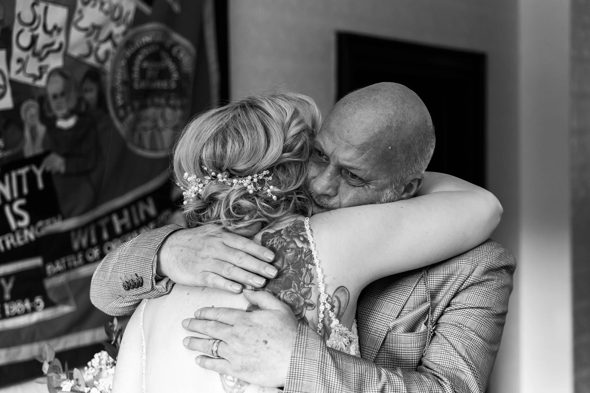 A bald groom in a checked suit warmly embraces a bride with shoulder tattoos in a tender, black and white moment.