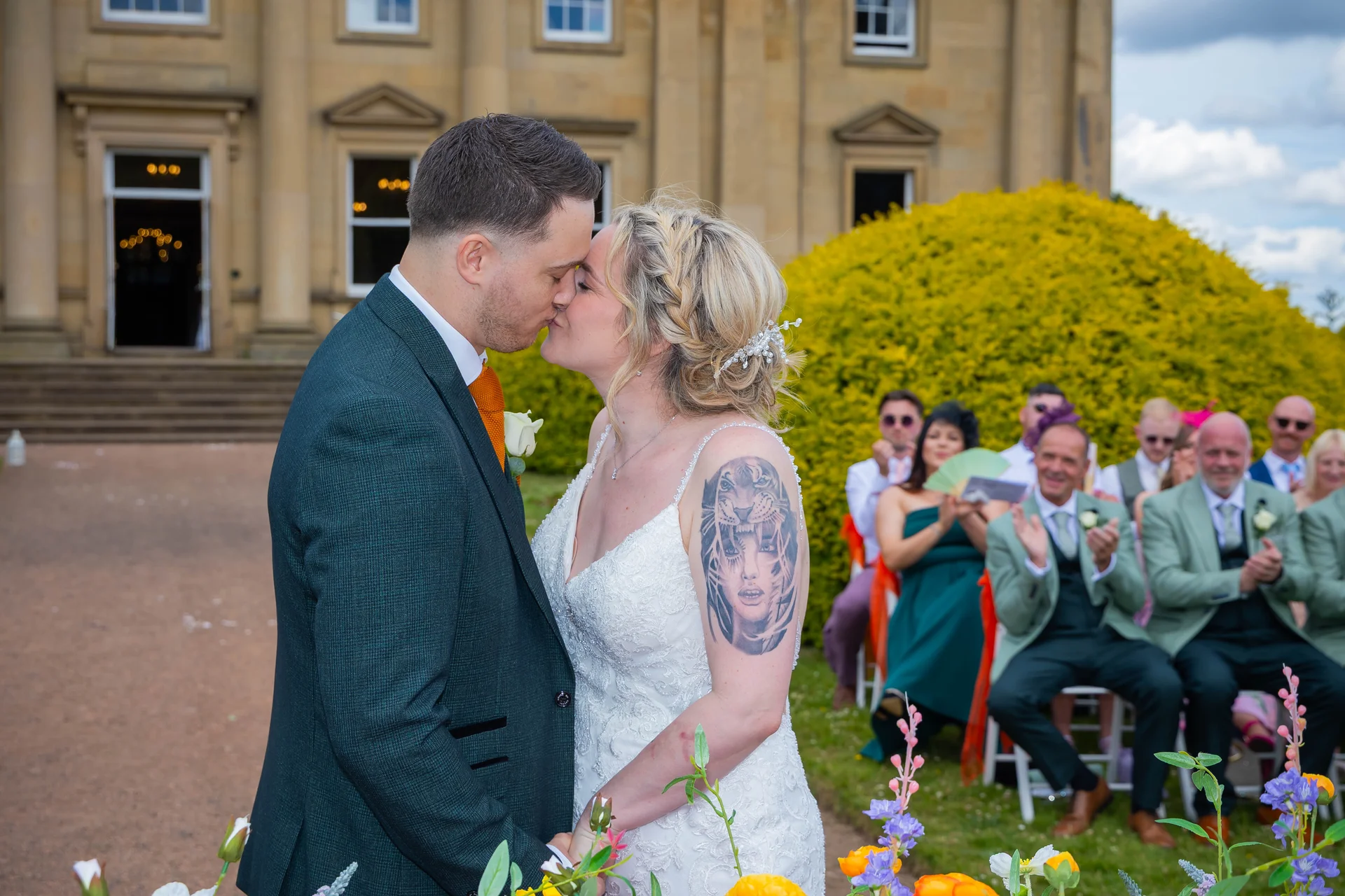 Newlywed bride and groom share their first kiss outdoors in front of a grand country house while seated guests clap and celebrate.