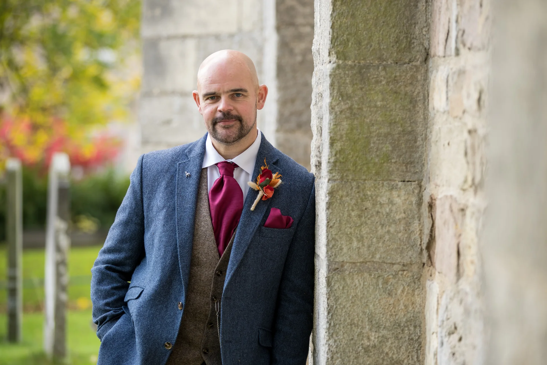 A solo groom in a blue tweed suit, brown waistcoat, and burgundy tie leans against an old stone wall outside a church.