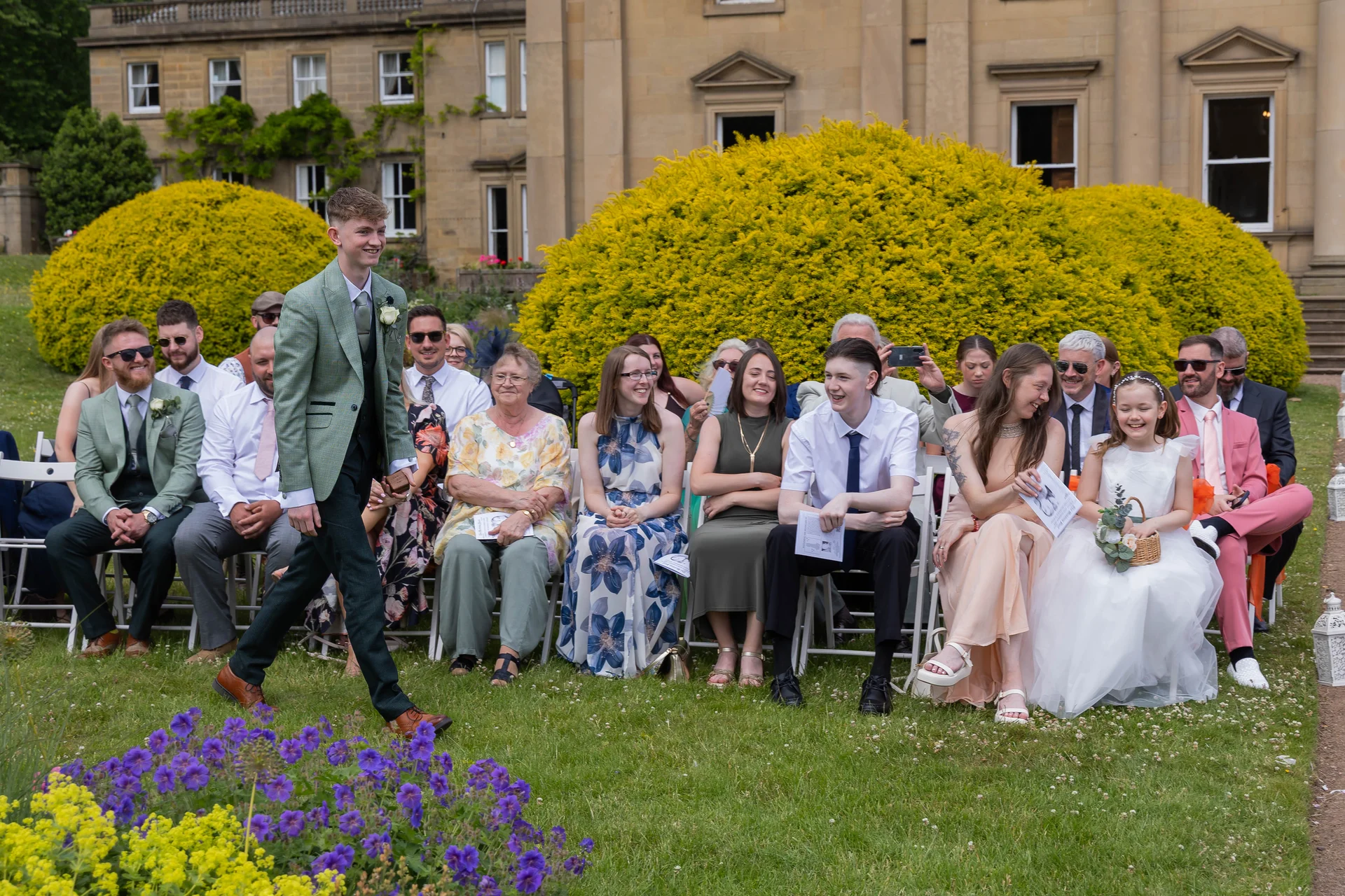 A young page boy in a green jacket walks happily past rows of seated wedding guests enjoying an outdoor ceremony on the lawn of a grand country house.