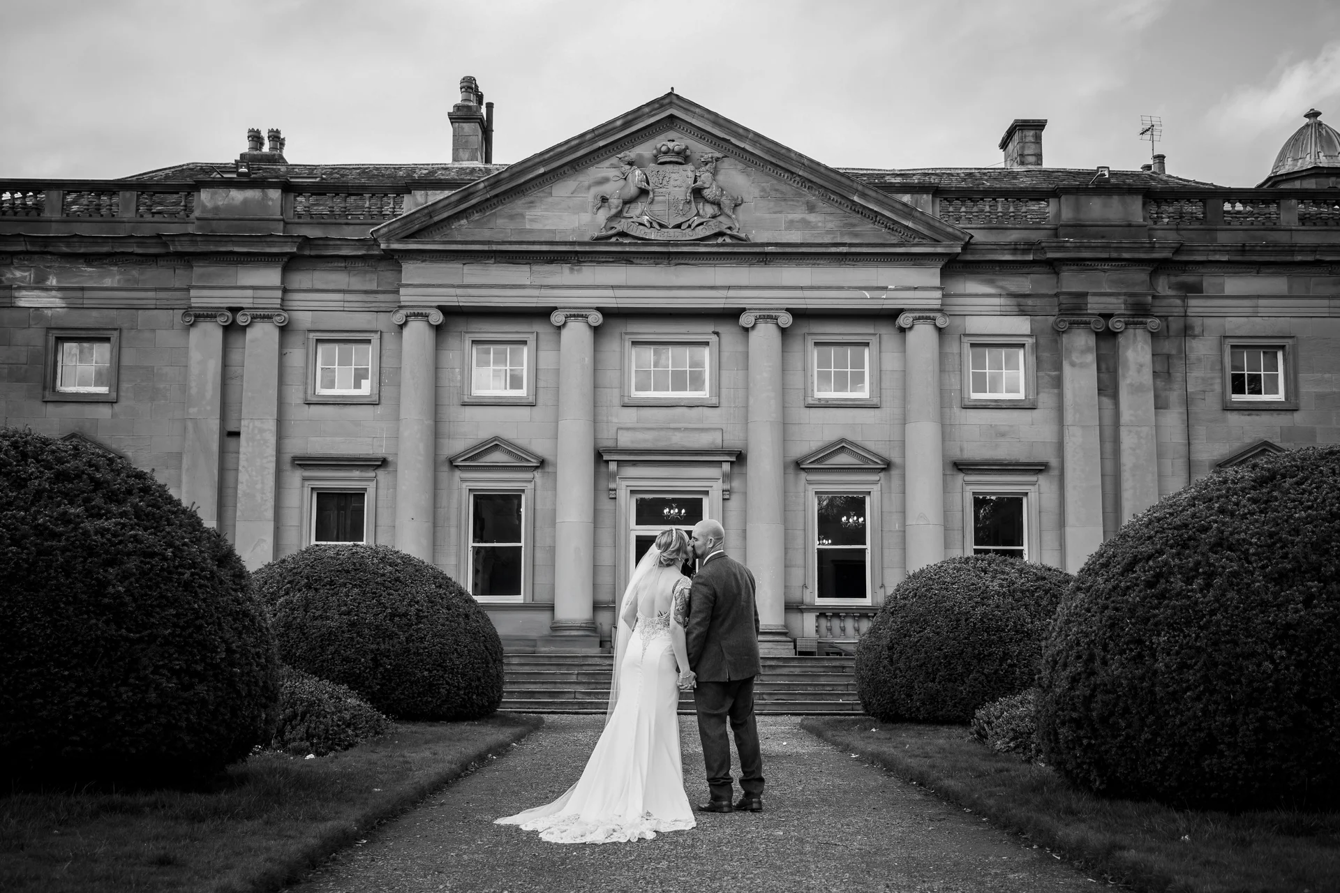 Newlyweds share a romantic kiss on the gravel path outside a grand stately home flanked by manicured topiary bushes.