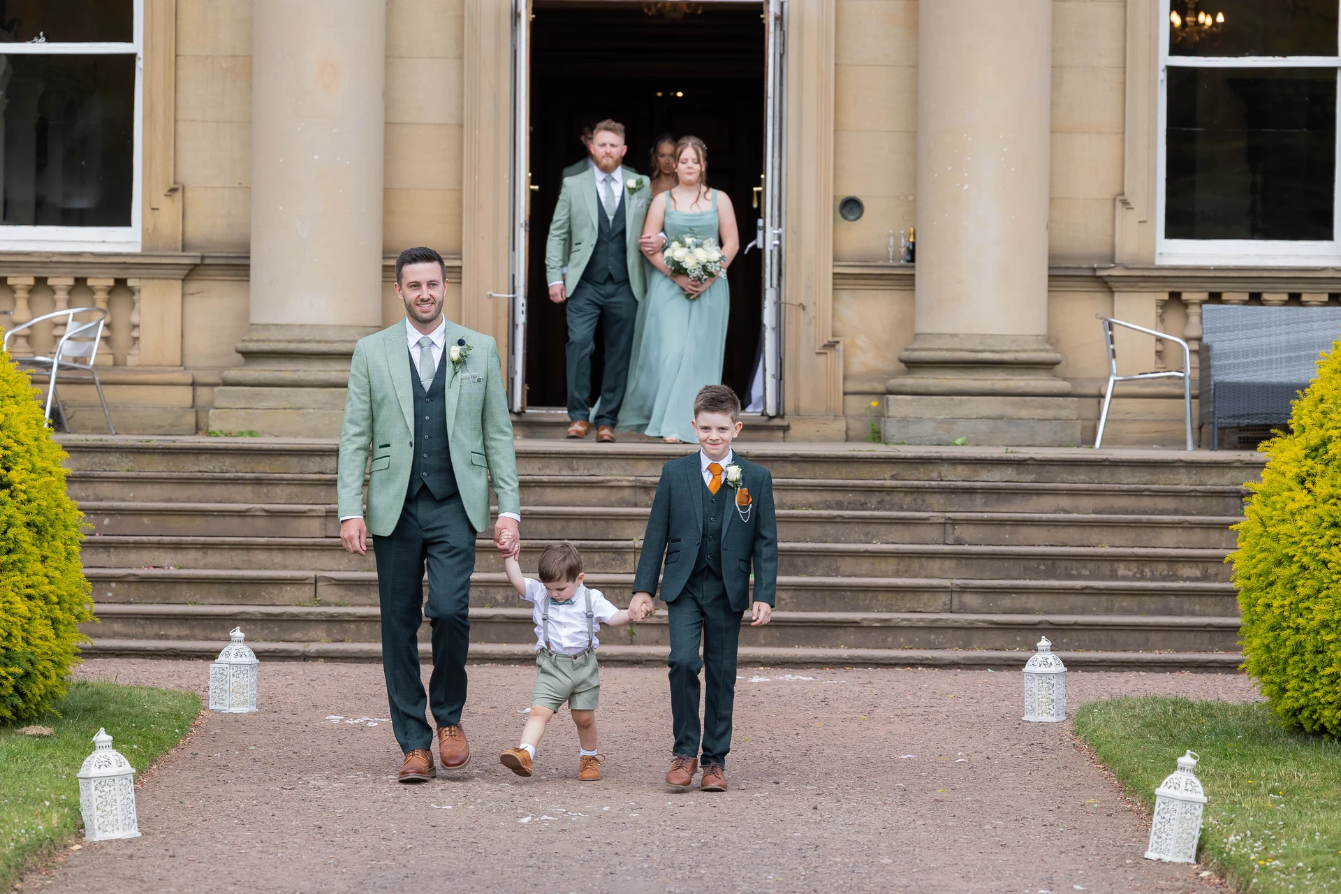A groom and young attendants walk hand in hand down stone steps outside a grand country house venue while the wedding party emerges from the doorway behind them.