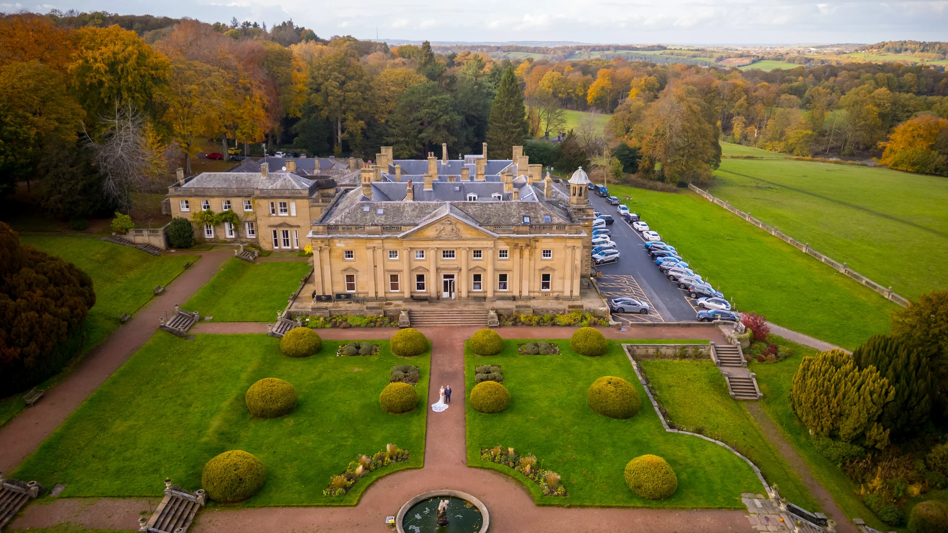 An aerial drone shot captures a newlywed couple standing on a pathway in the manicured formal gardens of a grand country house during autumn, surrounded by vibrant golden foliage and rolling countryside.