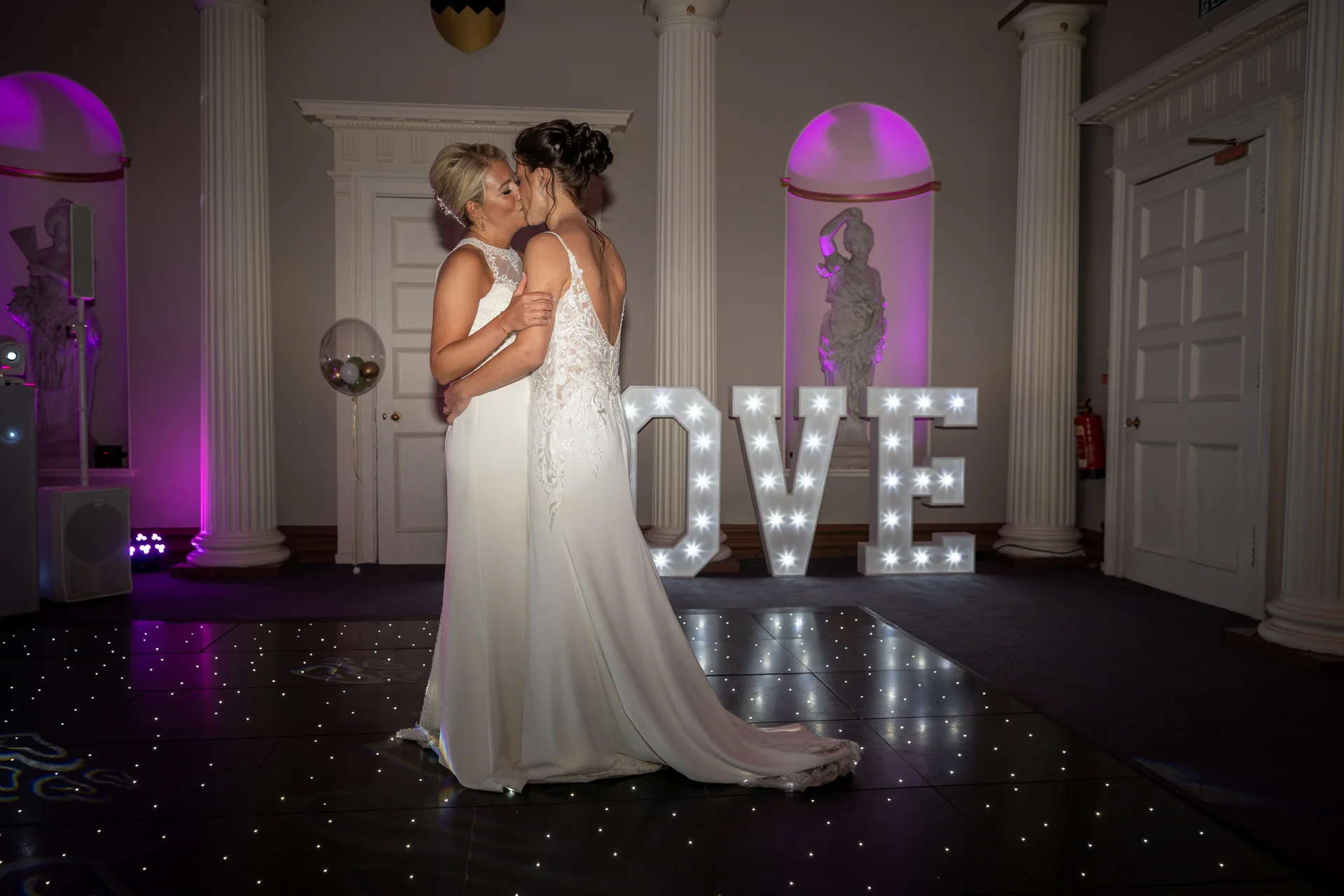 Two brides share a tender kiss on a glowing dance floor during their elegant evening reception in a stately ballroom.