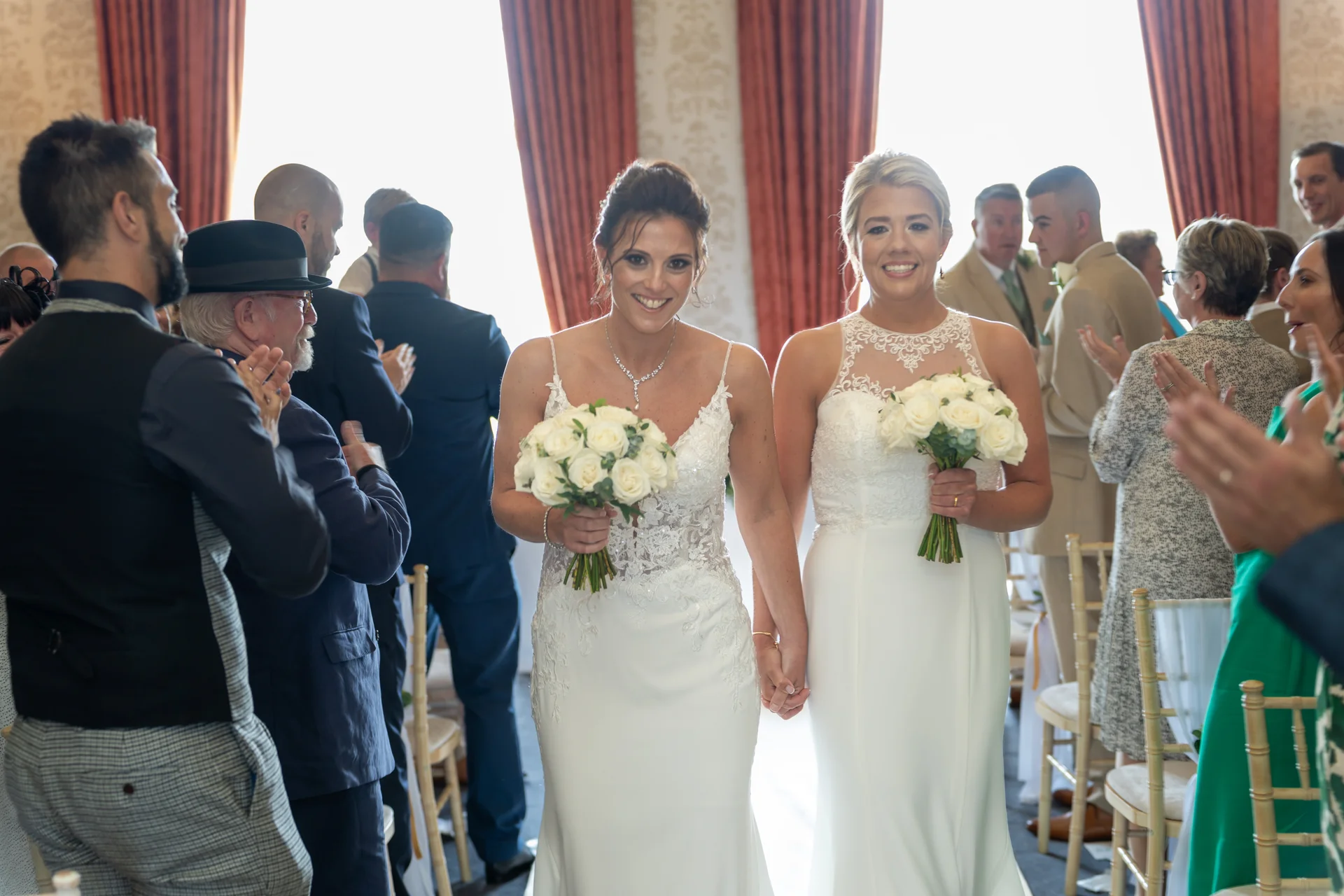 Two smiling brides holding hands and carrying white rose bouquets walk down the aisle as guests applaud and cheer.