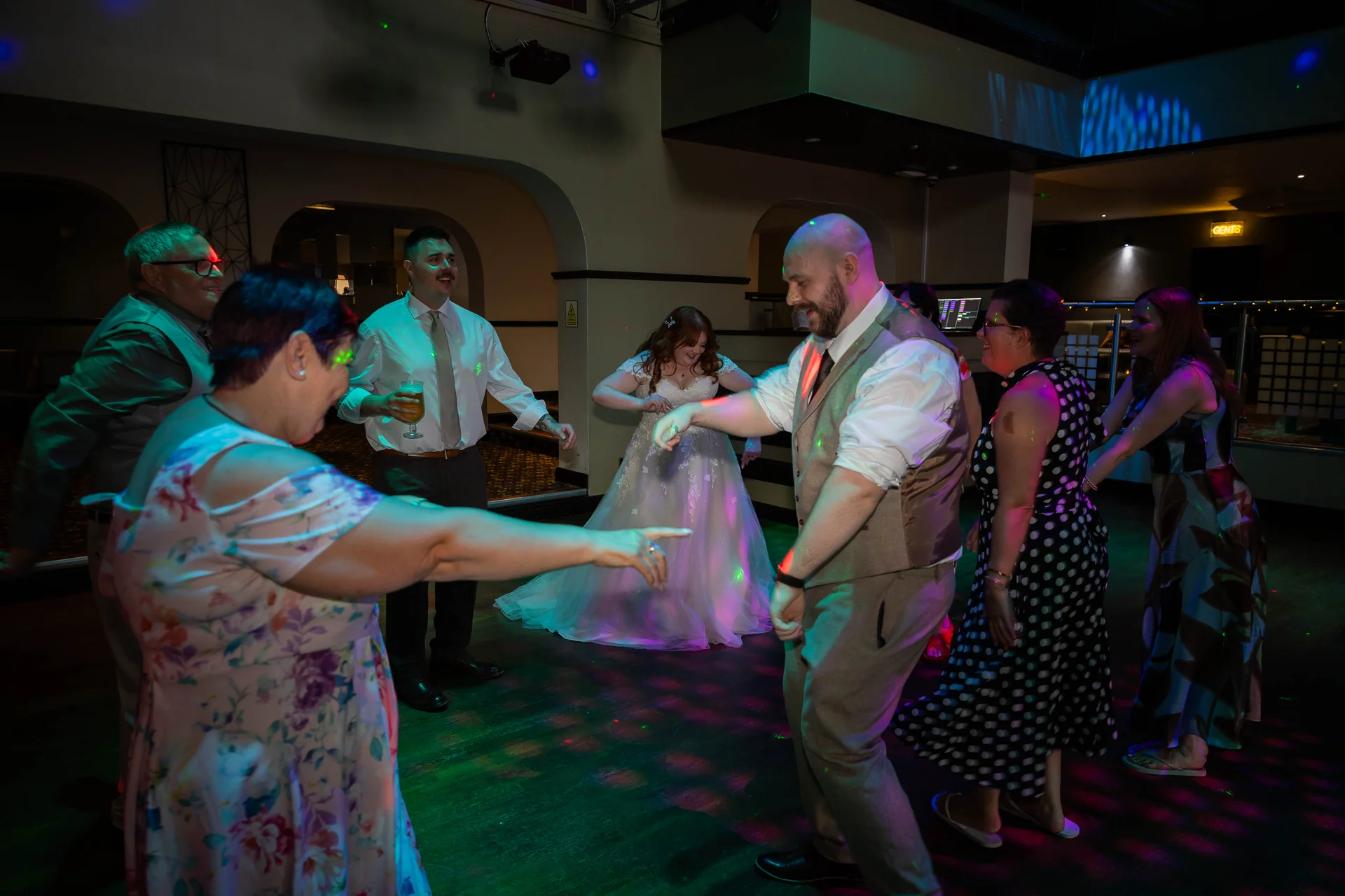 The bride and groom and guests enjoy dancing together on a lively illuminated dance floor during the evening wedding reception.