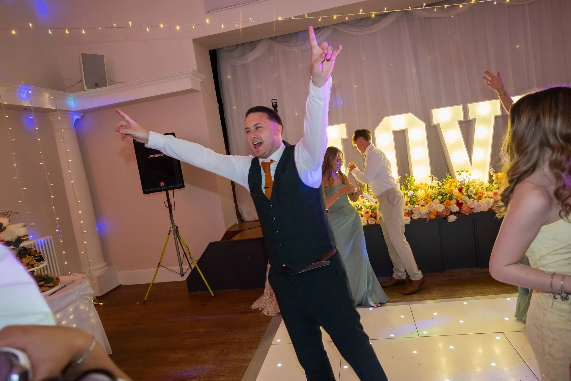 A joyful groom dances energetically with his arms raised high on an illuminated white dance floor during an evening wedding reception.