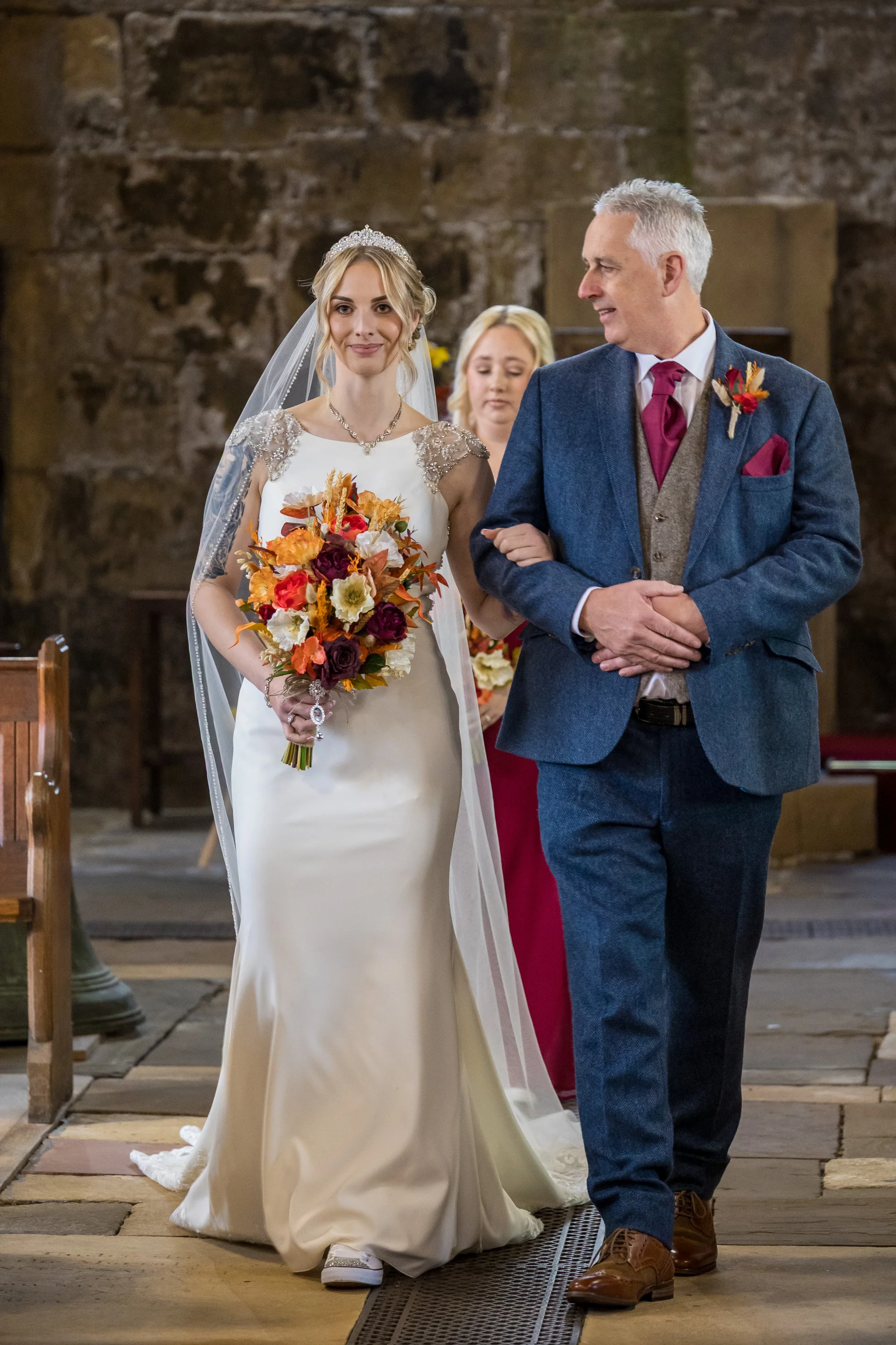 A smiling bride walking down the aisle of a historic stone church arm in arm with her father, holding a vibrant autumn bouquet and wearing a sleek white gown paired with trainers.