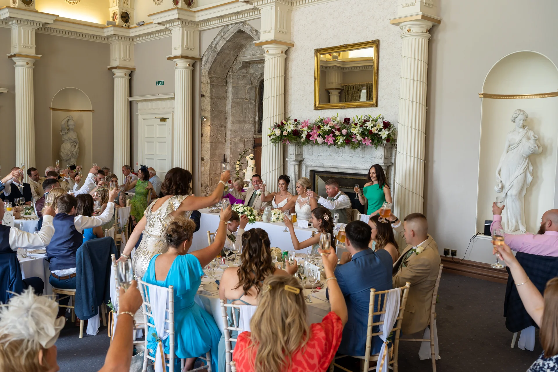Wedding guests raise their glasses for a toast during the wedding breakfast in a grand ballroom featuring classical columns and a fireplace adorned with floral arrangements.