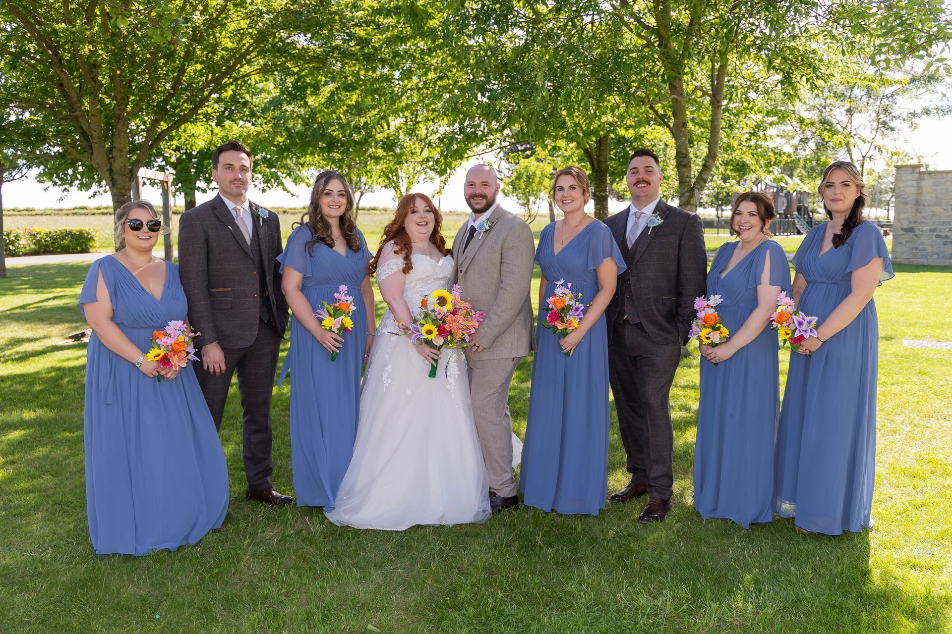 A smiling wedding party poses outdoors on a sunlit lawn beneath green trees, featuring a bride and groom surrounded by groomsmen in tweed suits and bridesmaids in long dusty blue dresses holding colourful bouquets.