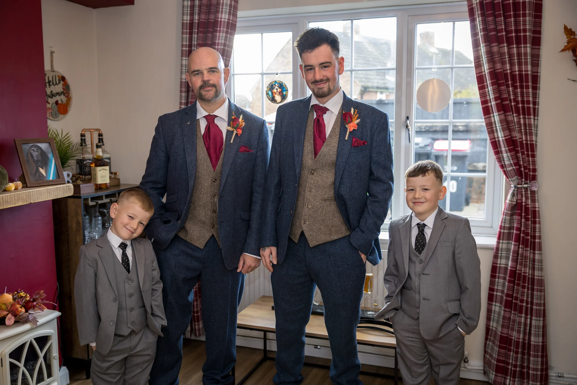 The groom and groomsmen stand together indoors wearing matching tweed suits with burgundy ties and pocket squares, accompanied by two young boys in grey suits.