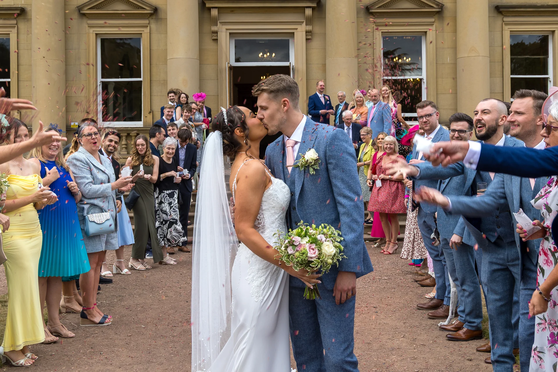 Newlyweds share a romantic kiss outside a grand country house as cheering wedding guests shower them with a flurry of colourful confetti.
