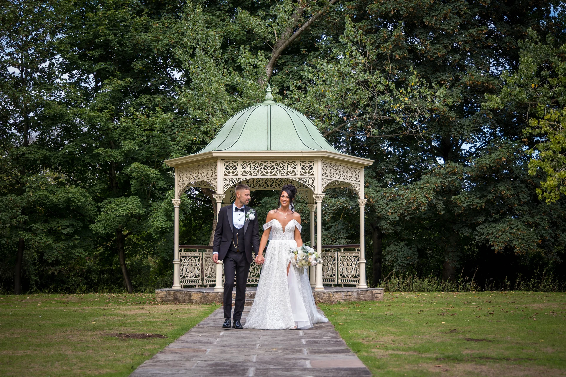 A newlywed couple walk hand in hand along a stone path in front of an ornate green and white bandstand surrounded by lush green trees.