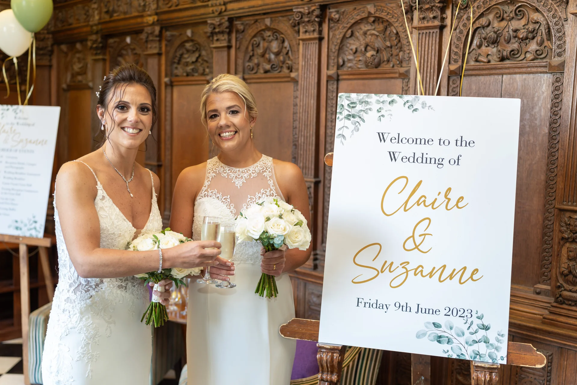 Two smiling brides in white wedding dresses hold bouquets and champagne flutes next to a welcome sign inside a wood-panelled room.