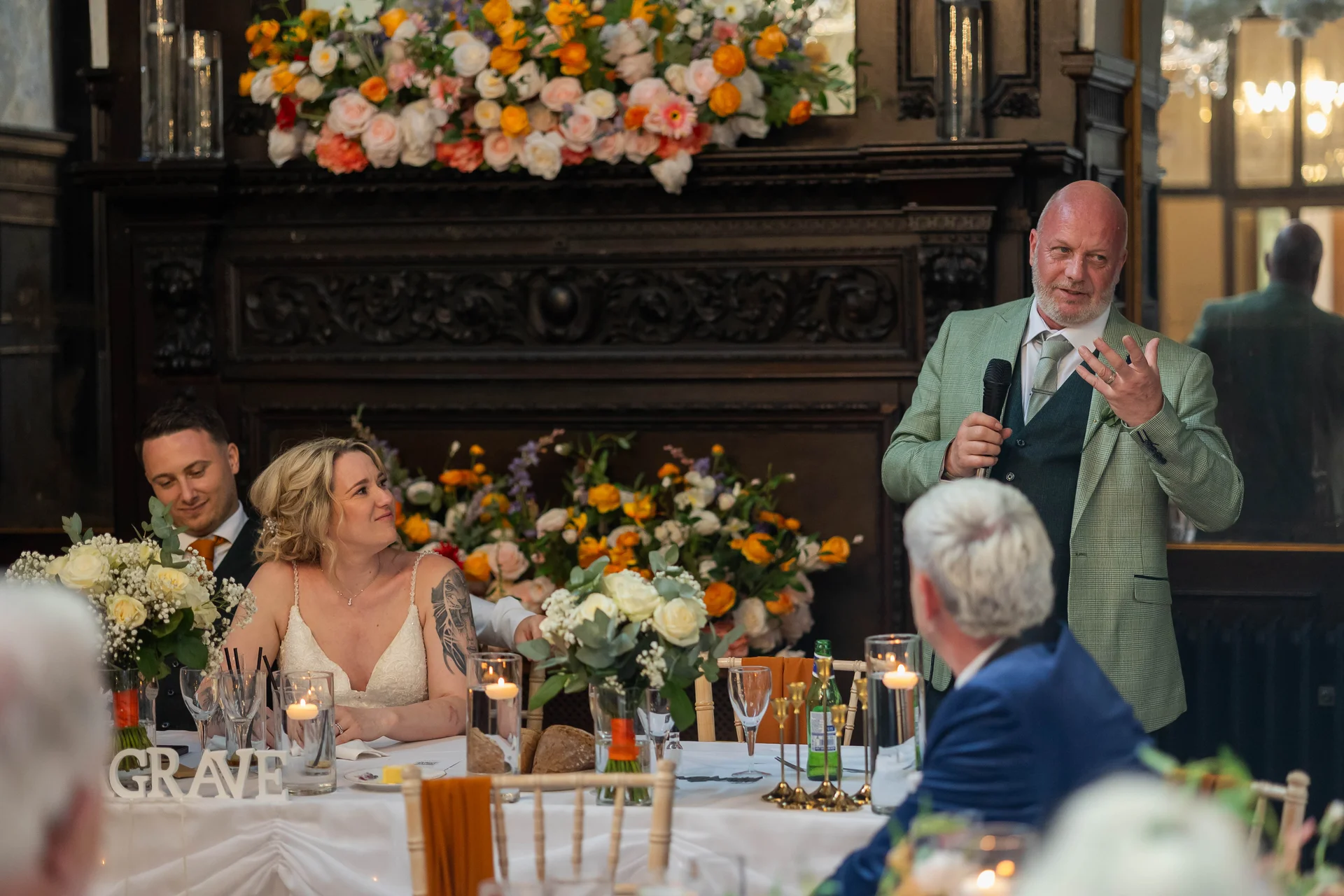 A guest delivers a speech holding a microphone at the top table, while the bride and groom look on smiling in a warmly lit, wood-panelled venue decorated with vibrant floral arrangements.