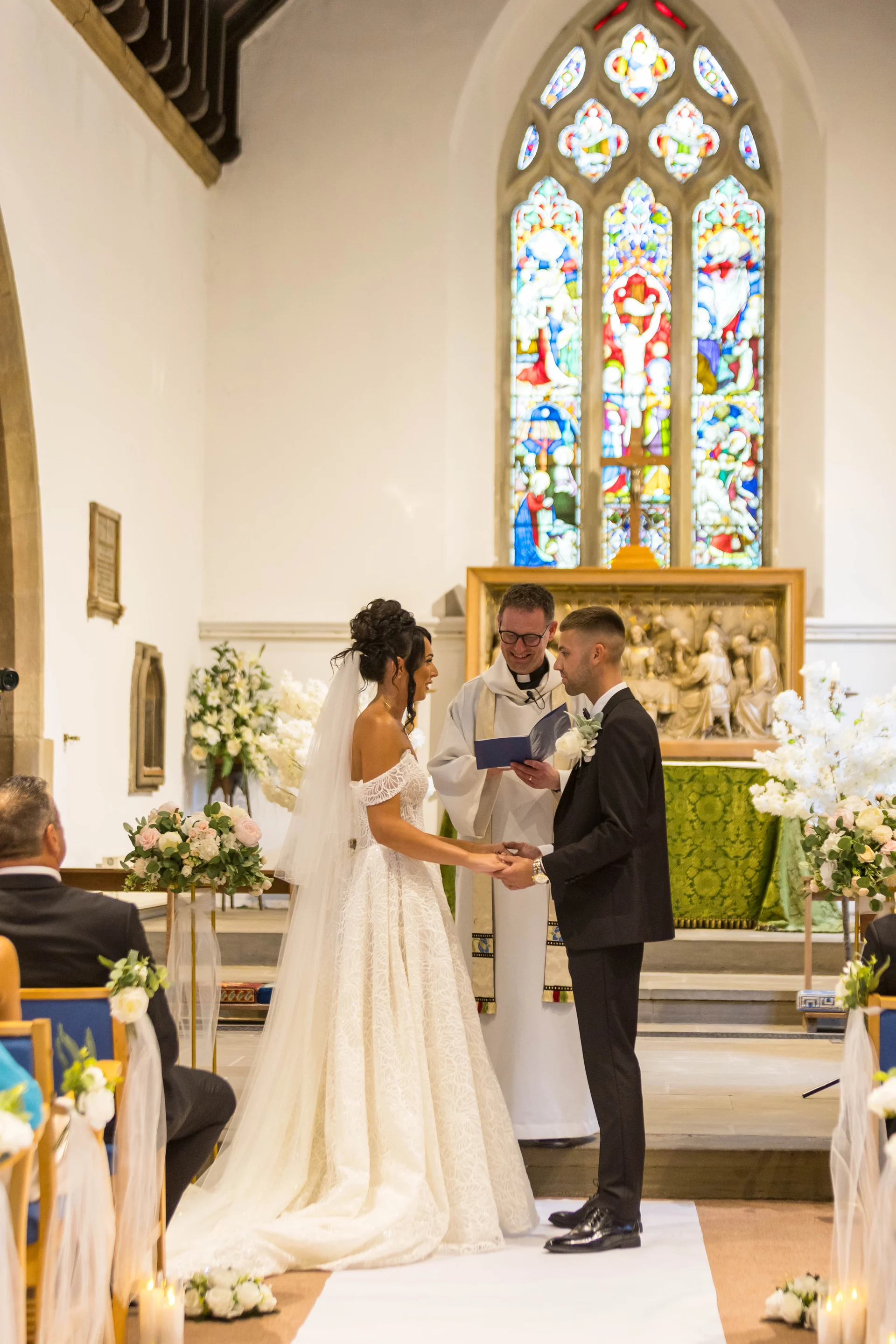 A bride and groom exchange vows at the altar of a traditional church with stunning stained glass windows, officiated by a robed vicar.