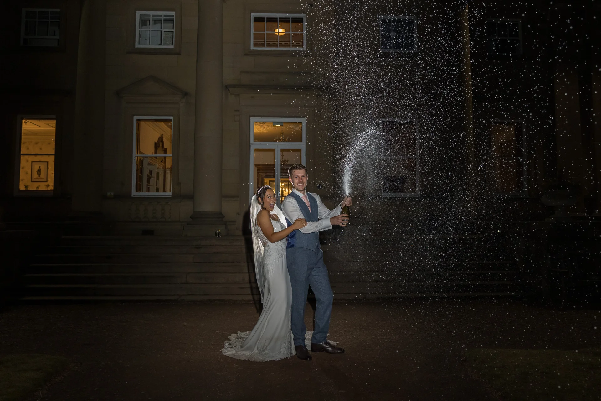A newlywed couple excitedly sprays champagne outside a grand manor house at night, illuminated by a dramatic flash against the dark sky.