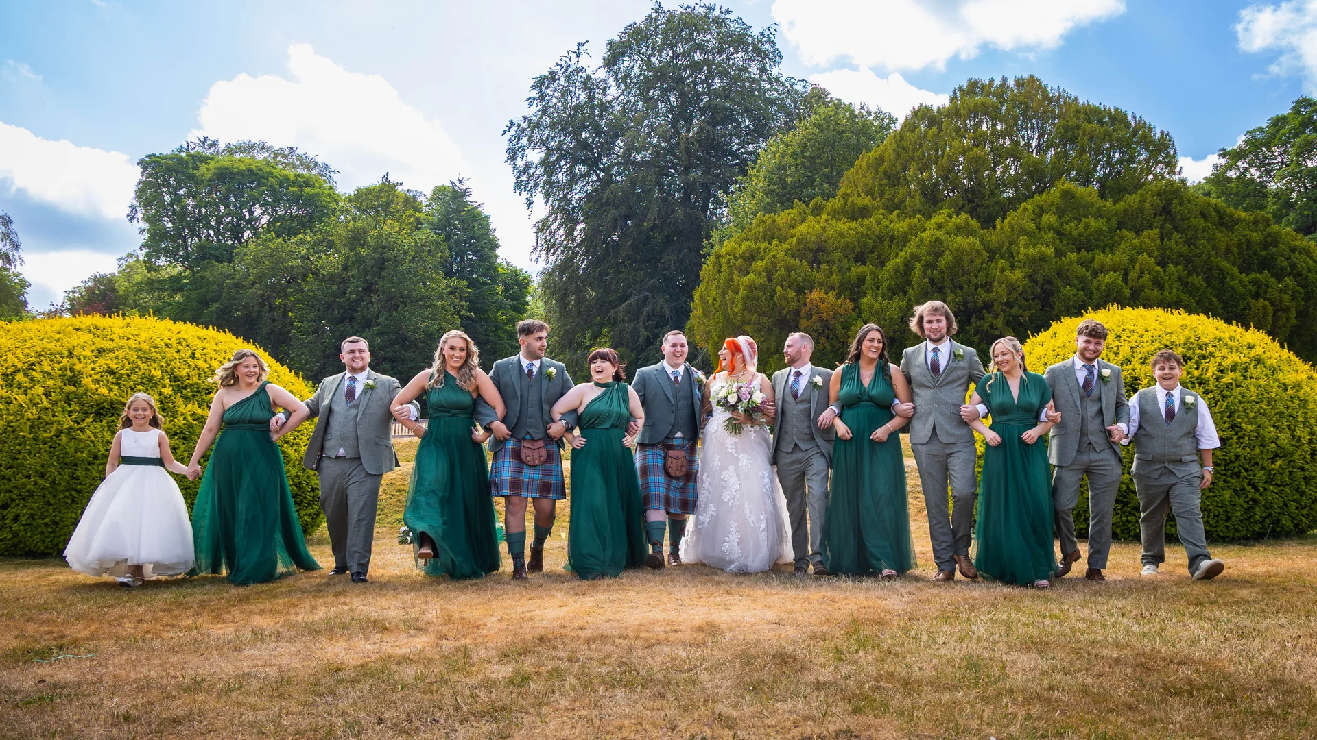 A large wedding party dressed in traditional Scottish kilts and vibrant green gowns walk hand in hand across a sunlit lawn lined with manicured hedges.