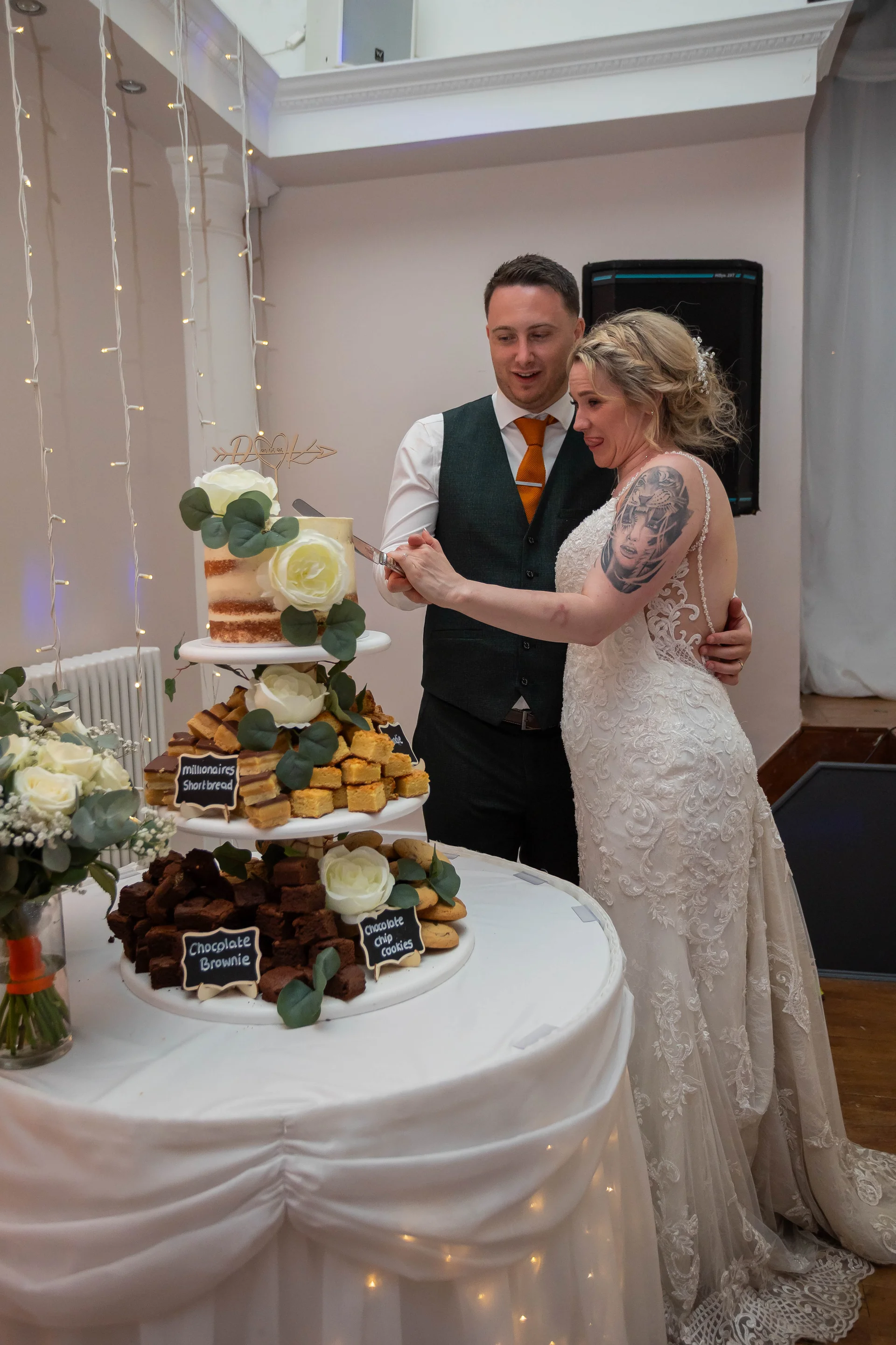 A bride and groom share a playful expression while cutting their multi-tiered wedding cake adorned with brownies and white roses.