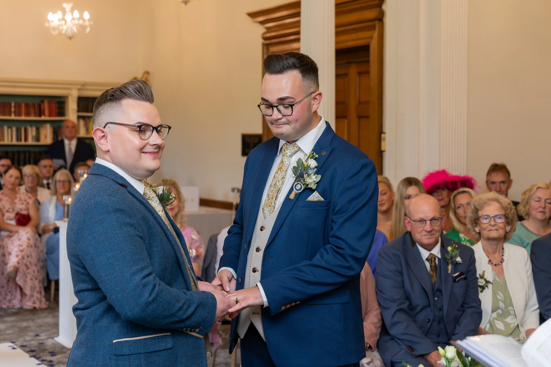 Two grooms exchange rings during a heartfelt civil ceremony inside a traditional venue with seated guests watching on.