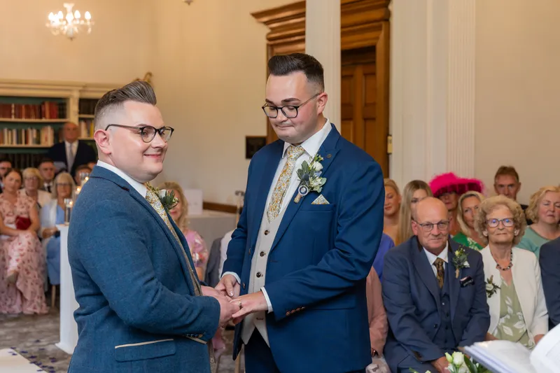 Two grooms exchange rings during a heartfelt civil ceremony inside a traditional venue with seated guests watching on.
