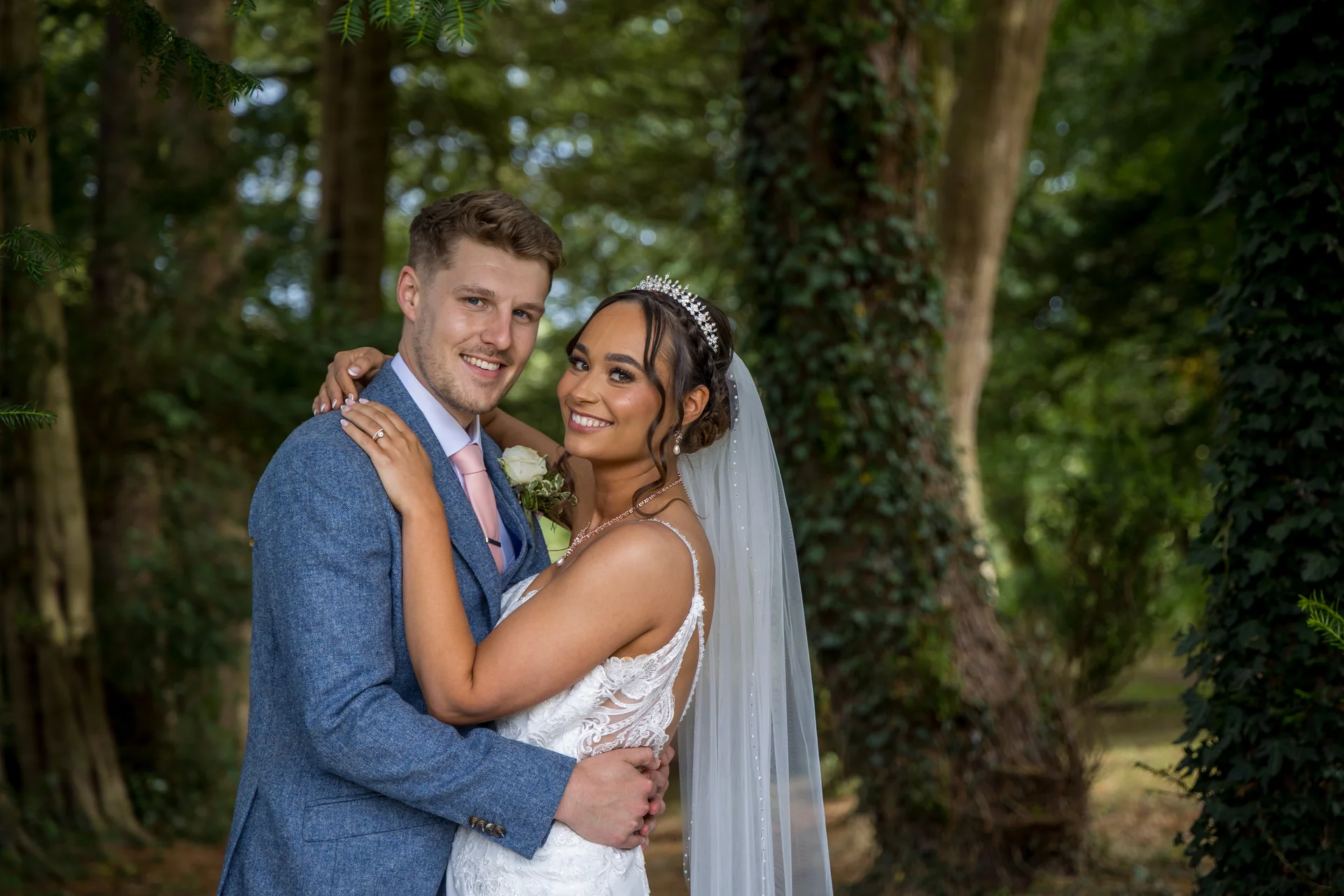 A smiling bride and groom embrace in a lush woodland setting, wearing a blue tweed suit and a lace wedding dress with a crystal tiara.