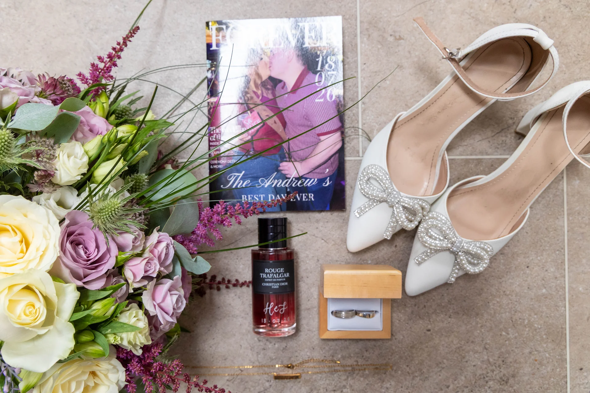 A bridal flat lay featuring a hand tied bouquet of roses and astilbe, white bow embellished heels, a perfume bottle, and wedding rings inside a wooden box on a tiled surface.
