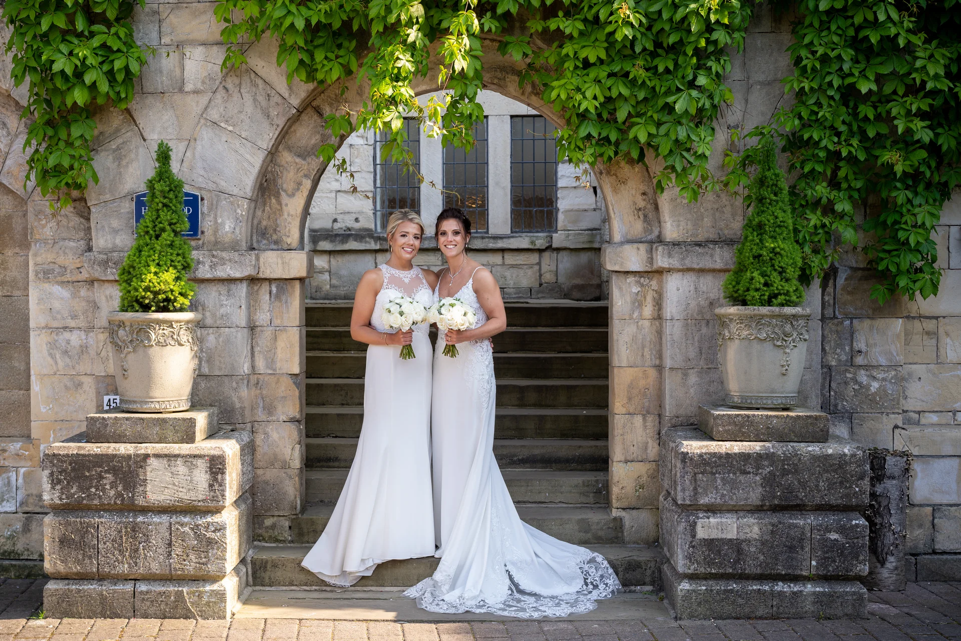 Two brides pose together holding white rose bouquets while standing in front of an ancient stone archway adorned with lush green ivy.