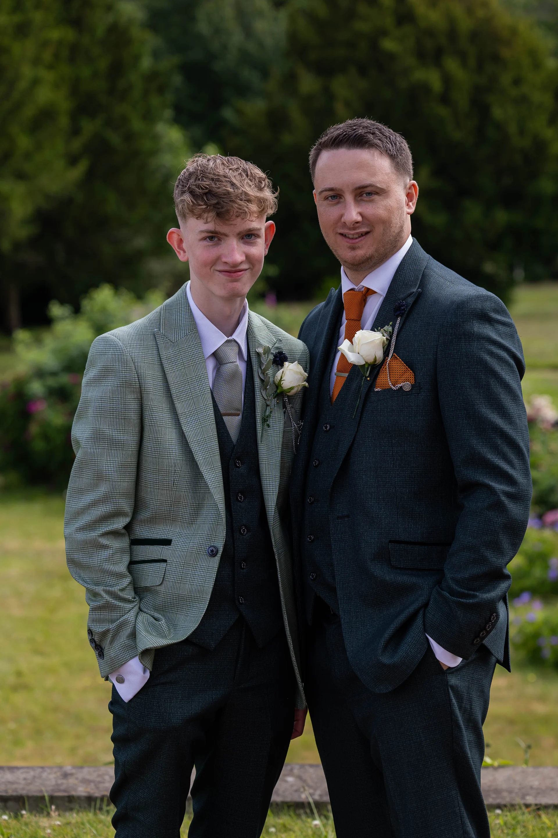 Two well-dressed men stand side by side outdoors wearing sophisticated three-piece suits with patterned jackets and matching floral buttonholes.