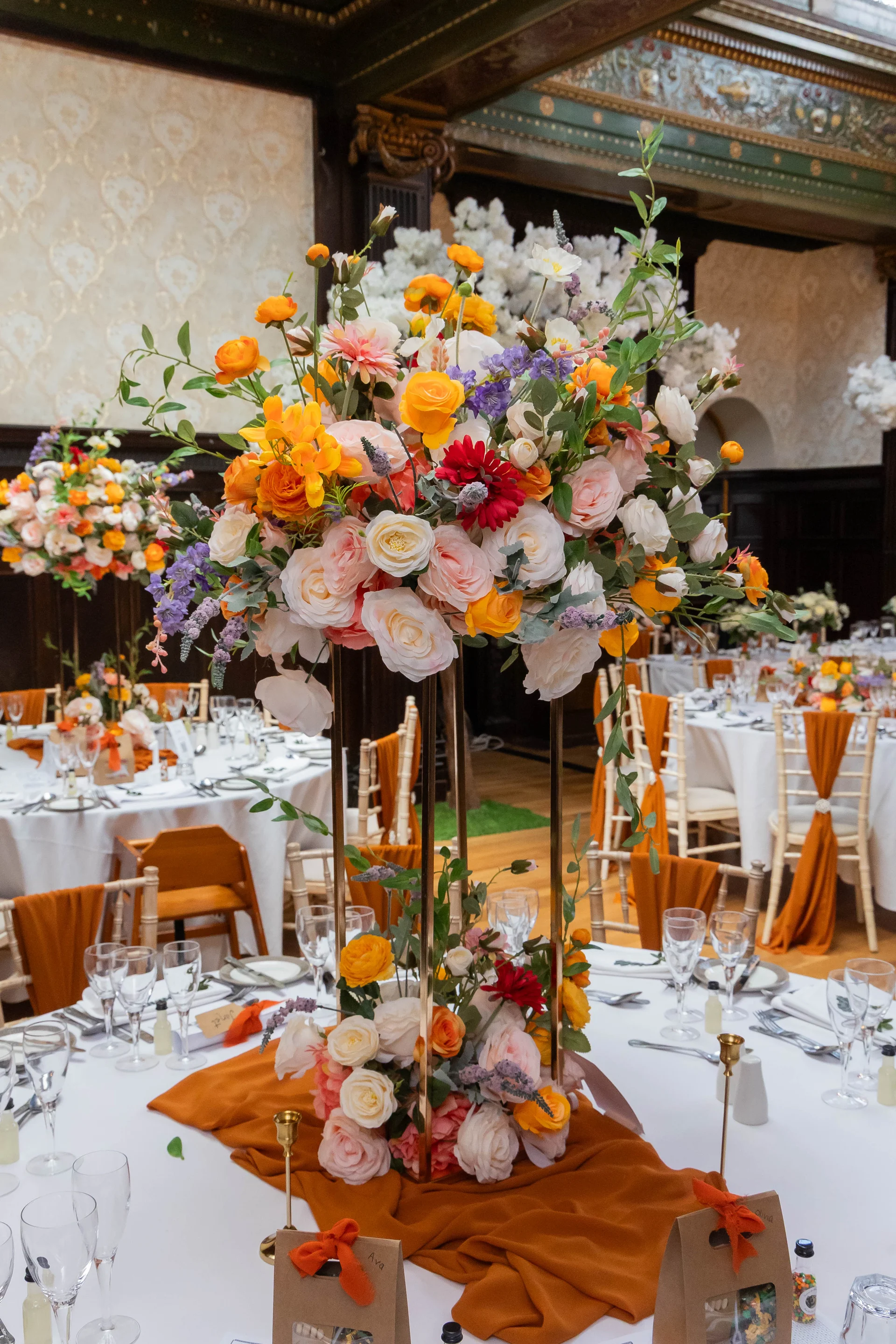 An elegant wedding reception room features round tables set with white linens, burnt orange runners, and tall floral centerpieces containing roses and ranunculus.