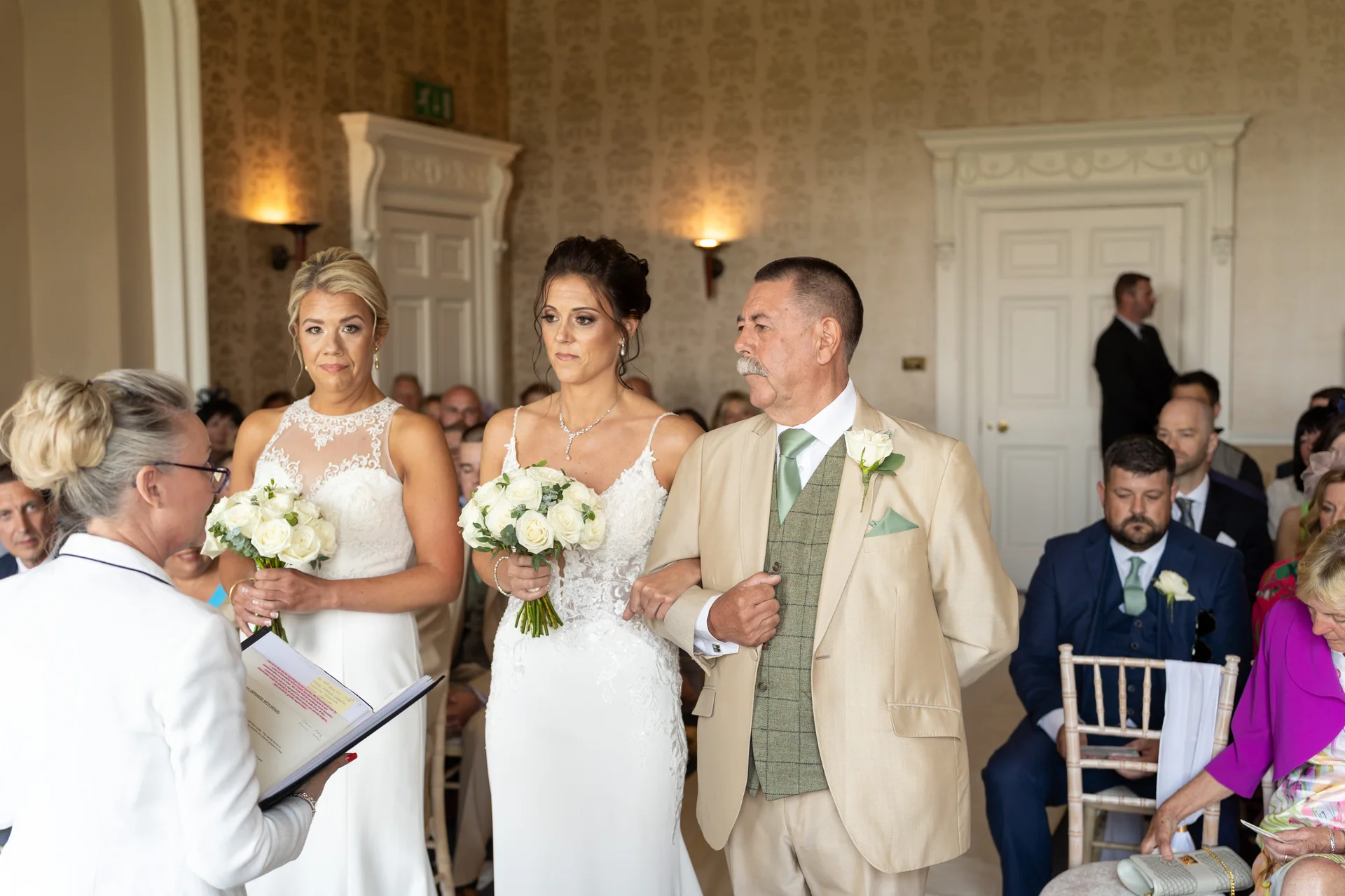 The bride stands arm in arm with her father in a light suit and waistcoat while a celebrant conducts the civil ceremony in an elegant room.