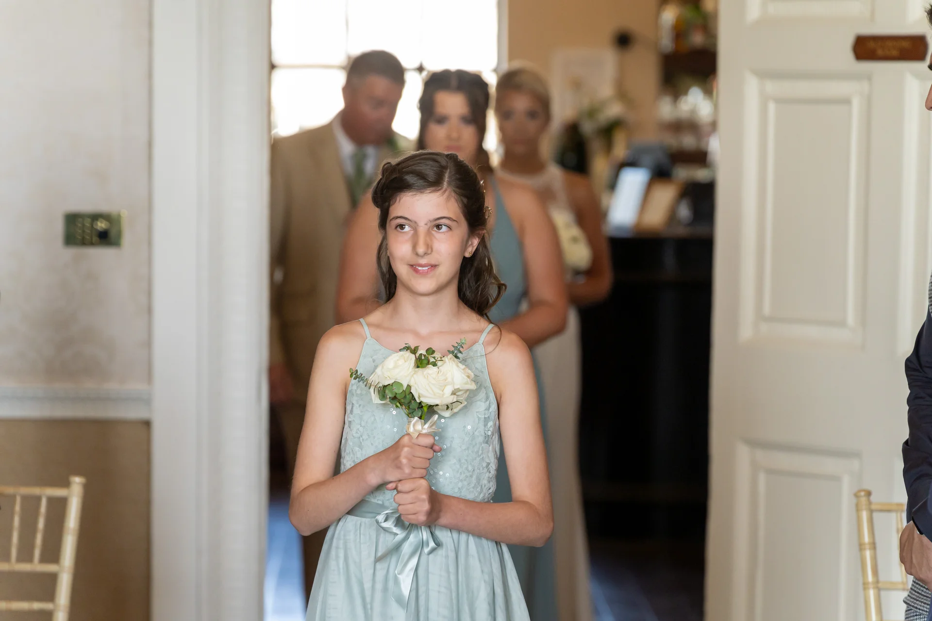 A young junior bridesmaid in a sage green dress holding a white rose posy walks down the aisle during a wedding processional.