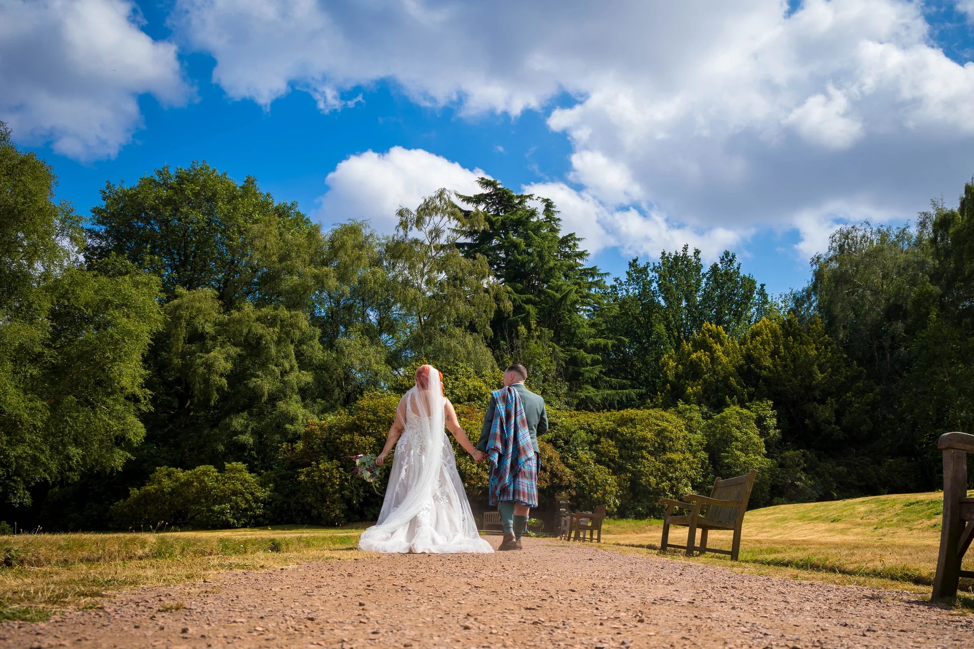 A newlywed couple walks hand in hand down a gravel path in a lush park, with the groom wearing a traditional tartan kilt and plaid sash.