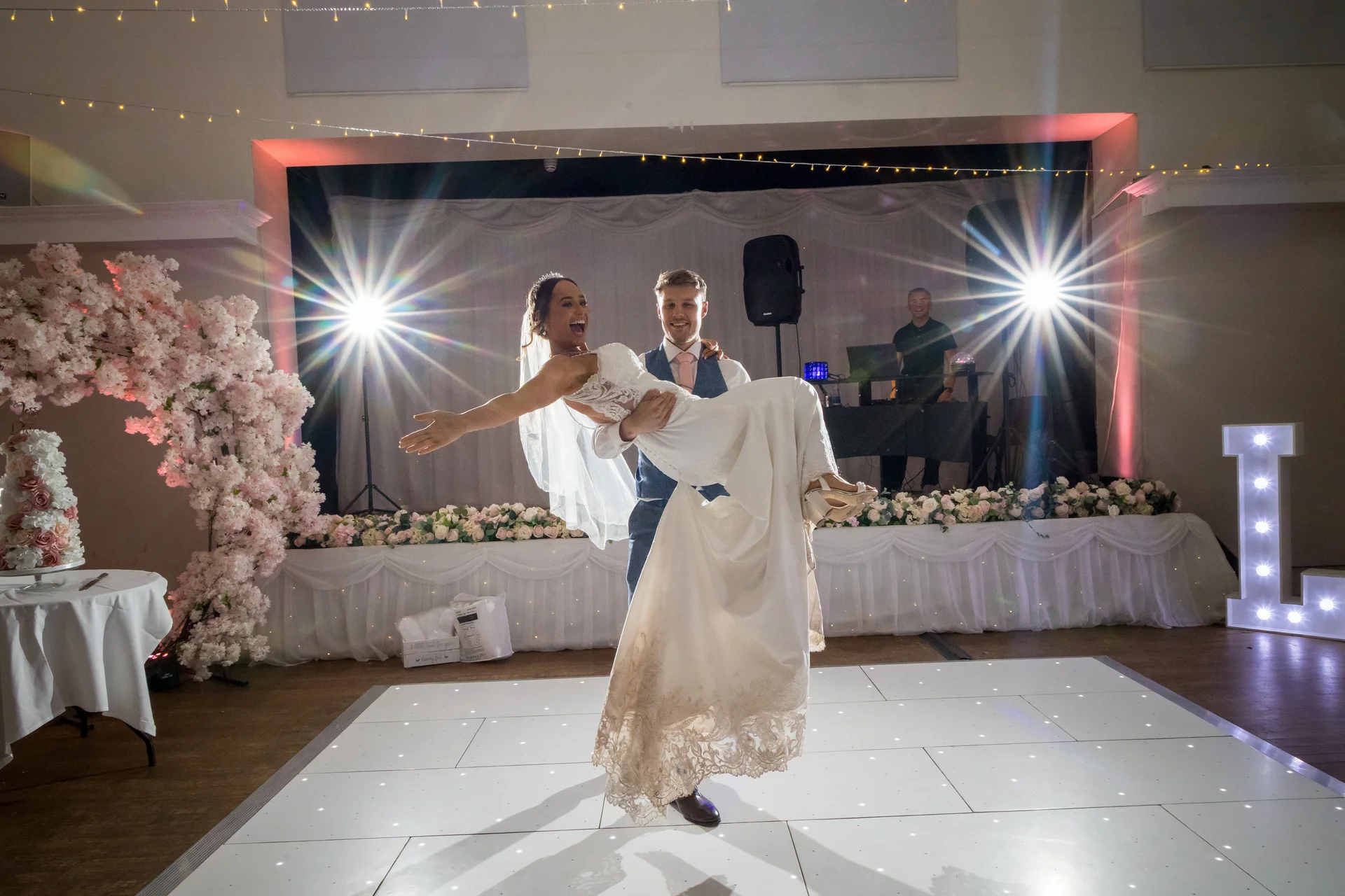 A groom joyfully lifts his laughing bride into his arms on a glowing white dance floor during their evening wedding reception.