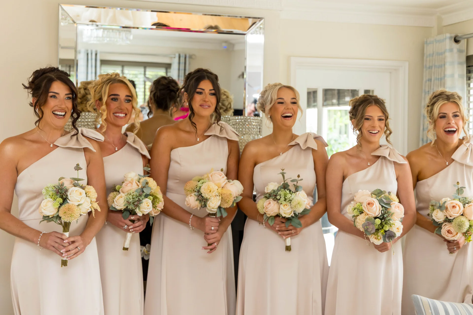 A line of six smiling bridesmaids in champagne one-shoulder satin dresses hold coordinating peach and cream rose bouquets indoors.