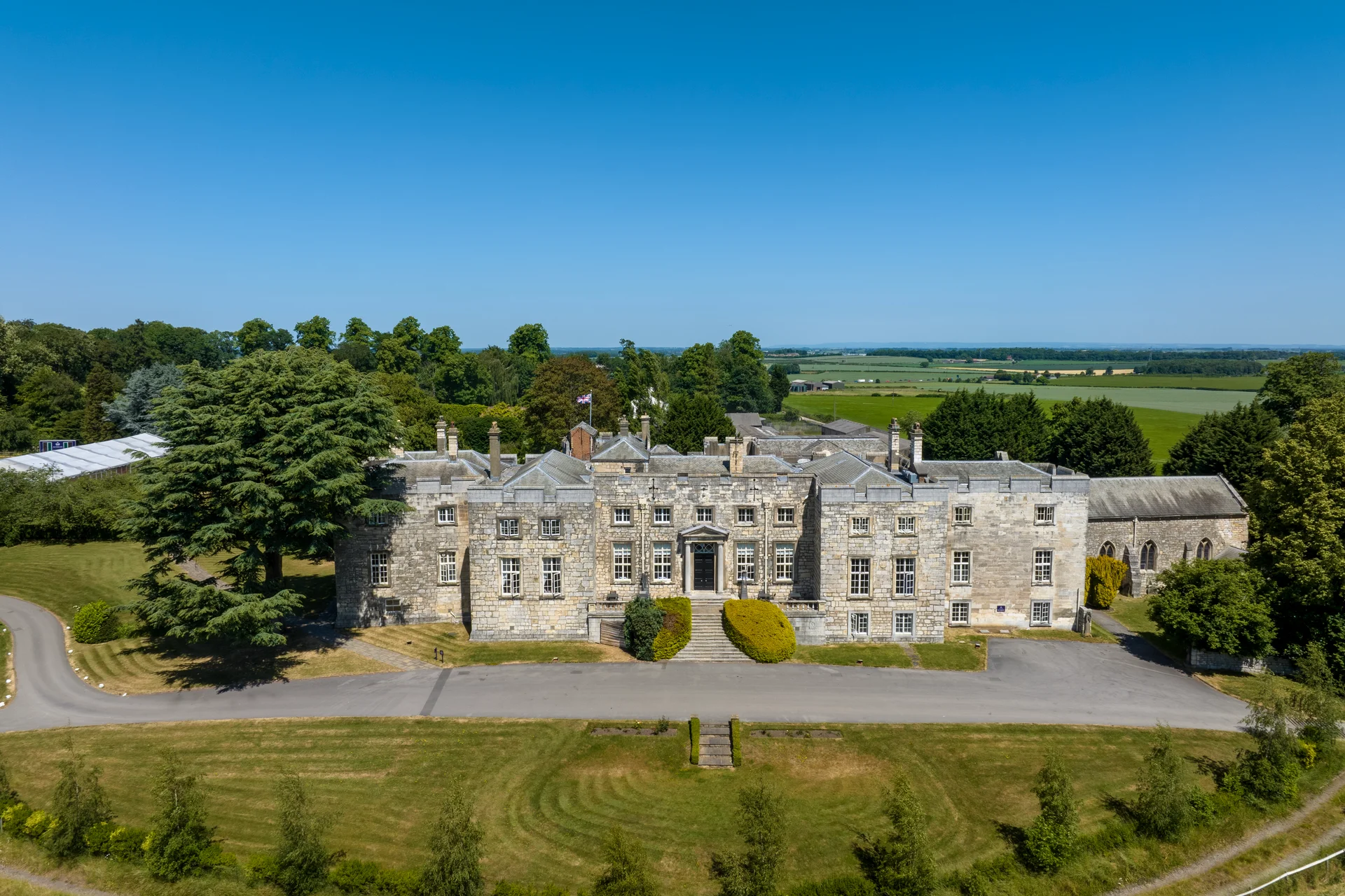 An aerial drone shot captures a grand stone country house wedding venue surrounded by manicured lawns, mature trees and sweeping countryside under a clear blue sky.