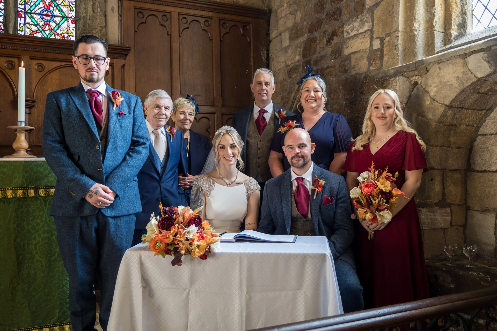 A bridal party and family members gather around the bride and groom as they sit signing the register inside a historic stone church.
