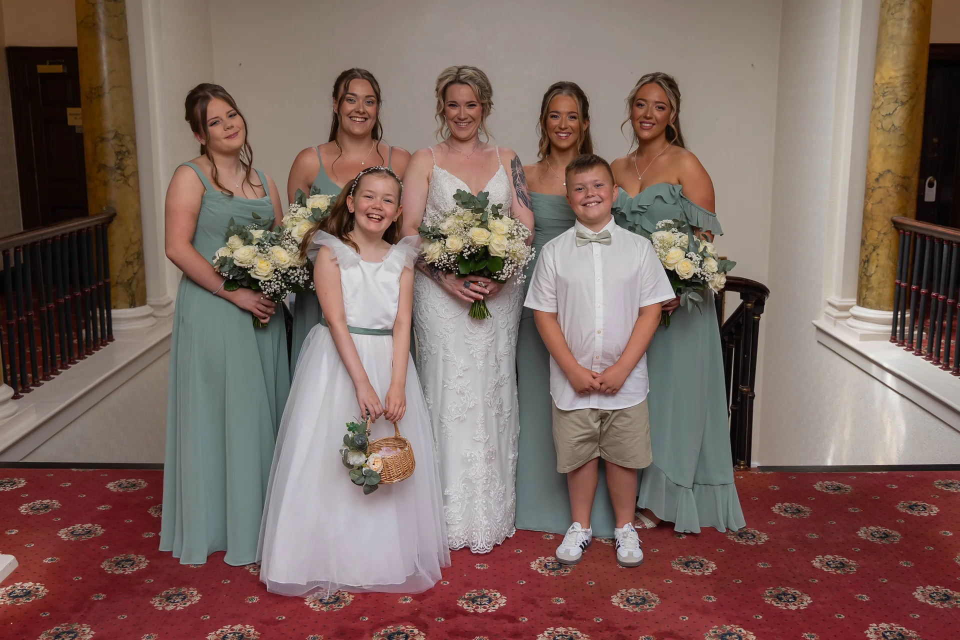 The radiant bride stands proudly in her lace wedding dress surrounded by her bridesmaids in sage green gowns, alongside a smiling flower girl and page boy holding a small wicker basket.