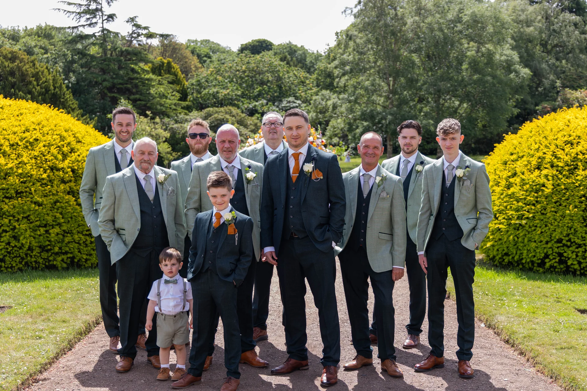 The groom and his groomsmen pose together on a gravel path outdoors on a sunny summer wedding day.