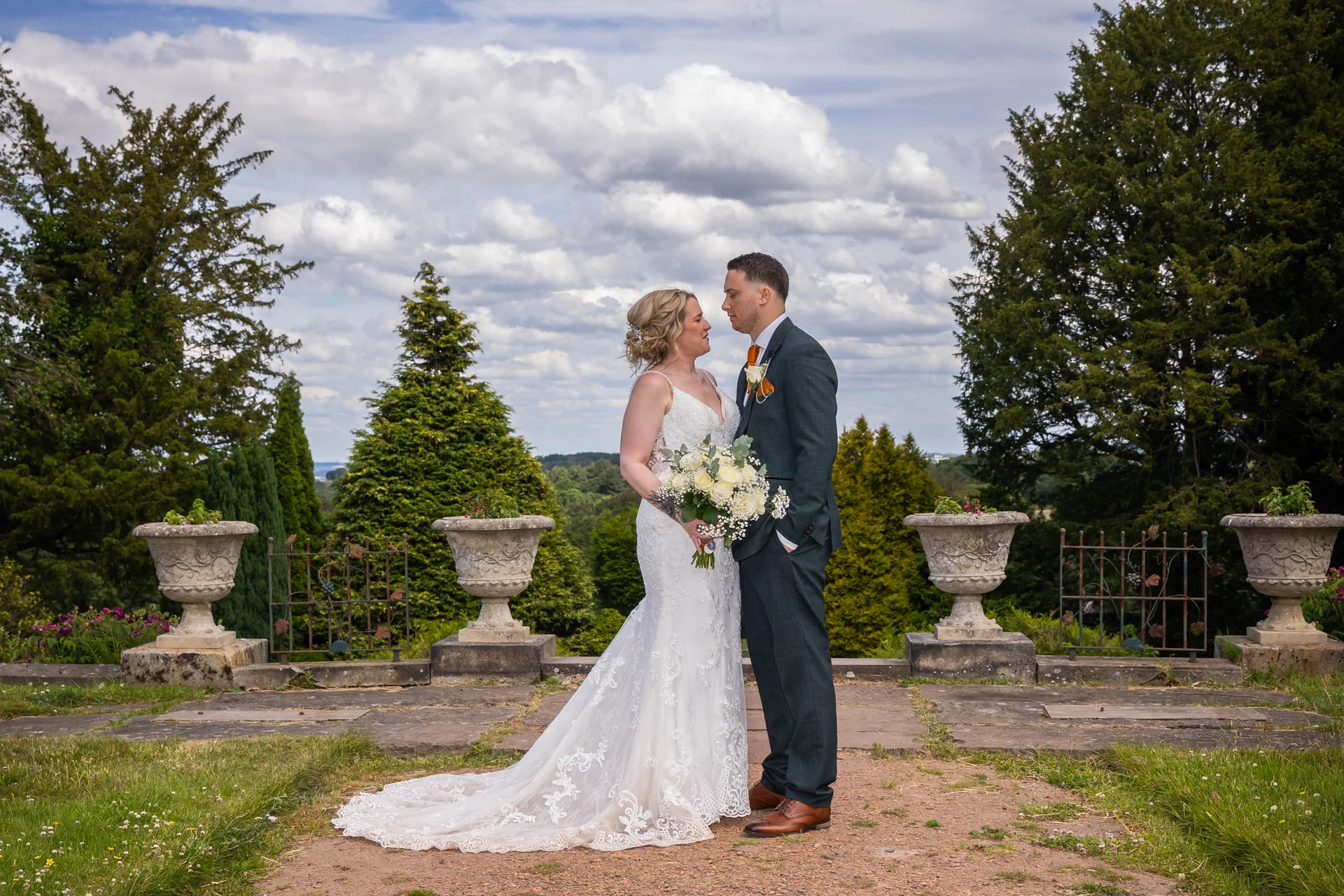 A newlywed couple stands face to face on a stone terrace overlooking rolling countryside beneath a dramatic cloudy sky, with the bride holding a bouquet of white roses.