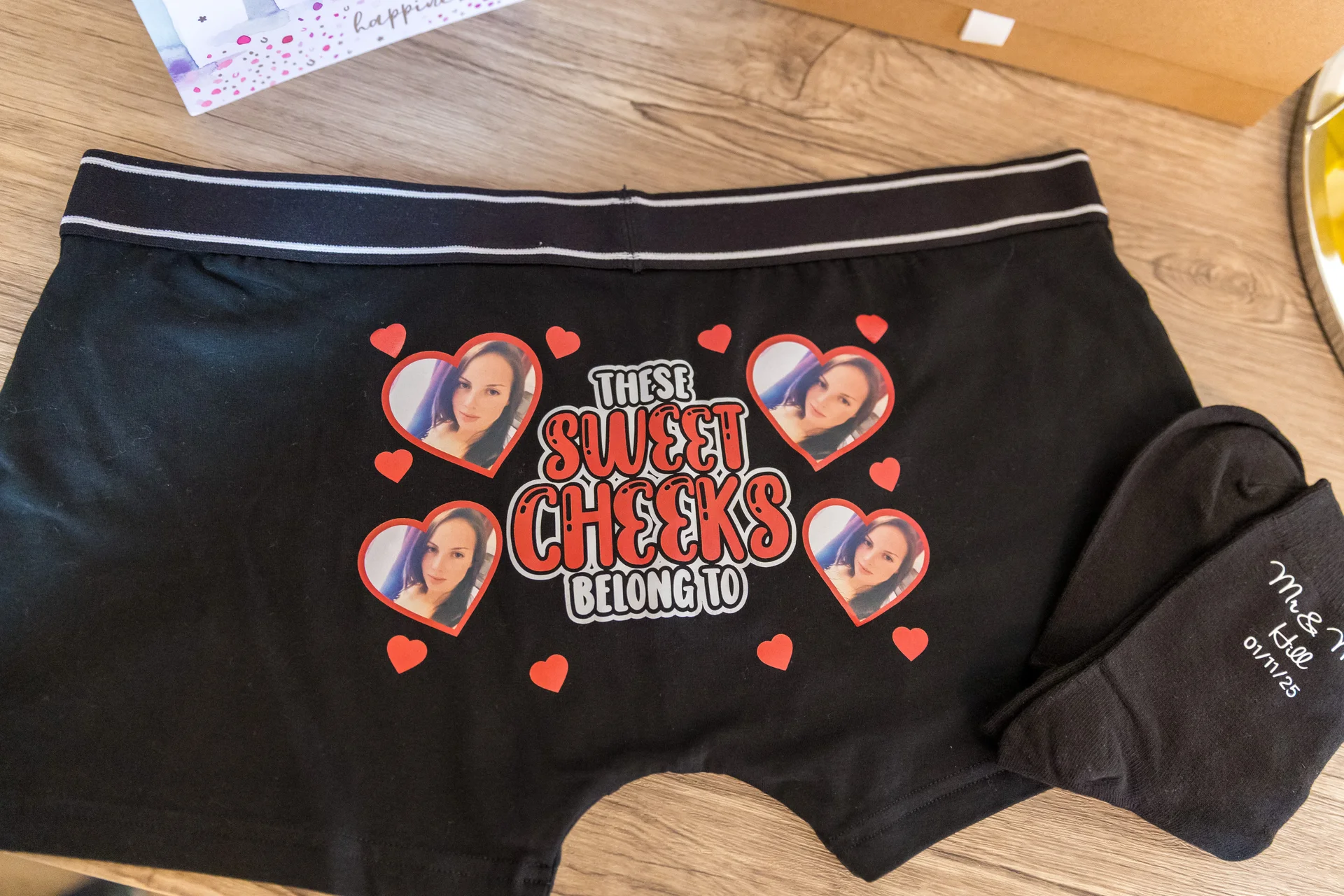 A flat lay image on a wooden surface shows novelty black groom underwear printed with a personalised face design and the text these sweet cheeks belong to, alongside matching black socks.