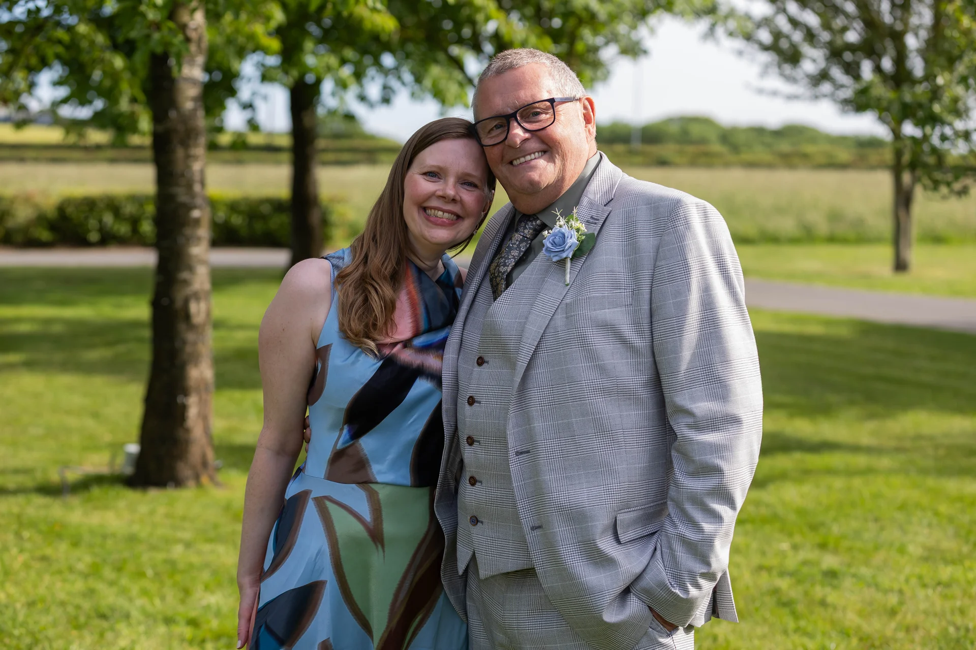 An older man in a light grey checked suit with a blue boutonniere stands smiling next to a smiling woman wearing a colourful patterned blue dress outdoors in a sunny garden.